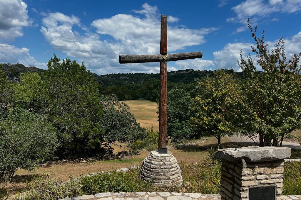Wooden cross overlooking a field and trees.