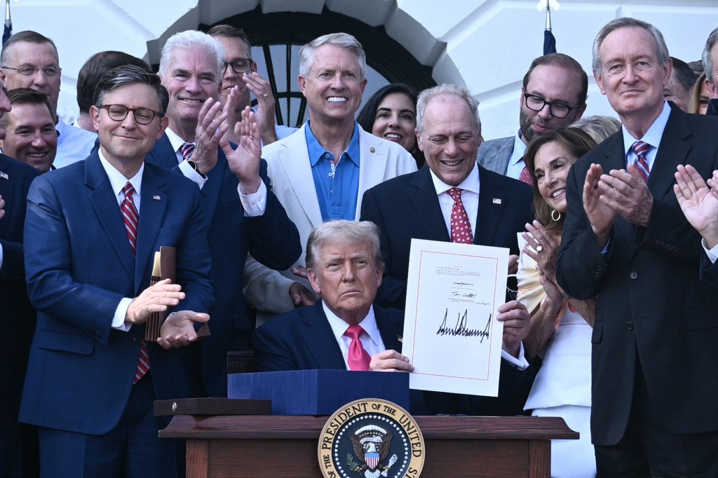 President Trump signing a bill at the White House, surrounded by applauding officials.