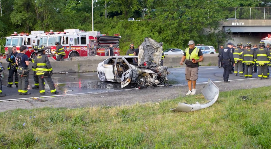 Aftermath of a multi-vehicle crash on a highway, showing a burned-out car and emergency responders.