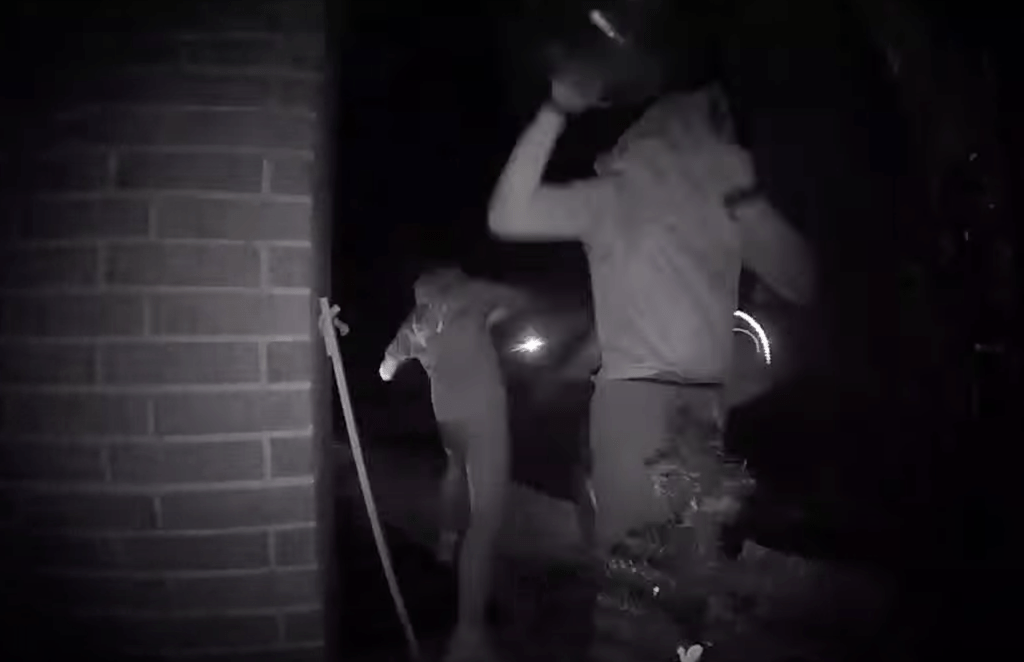 Night vision security camera footage of two hooded figures vandalizing property.