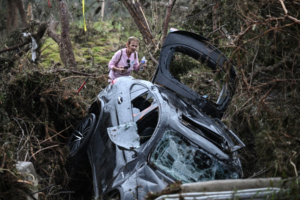 Woman holding a water bottle and phone near a damaged car in flood debris.