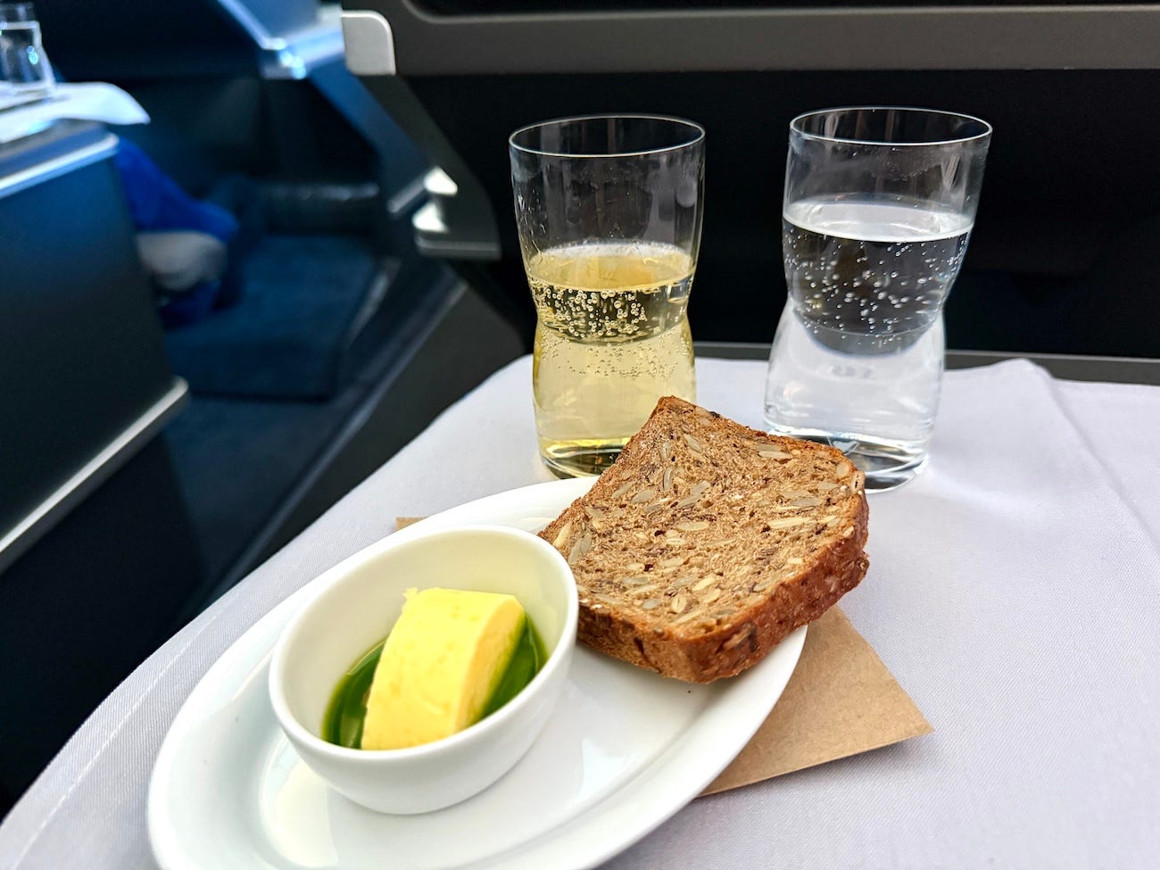 Slied of bread on plate next to bowl of butter with glass of water, champagne behind it