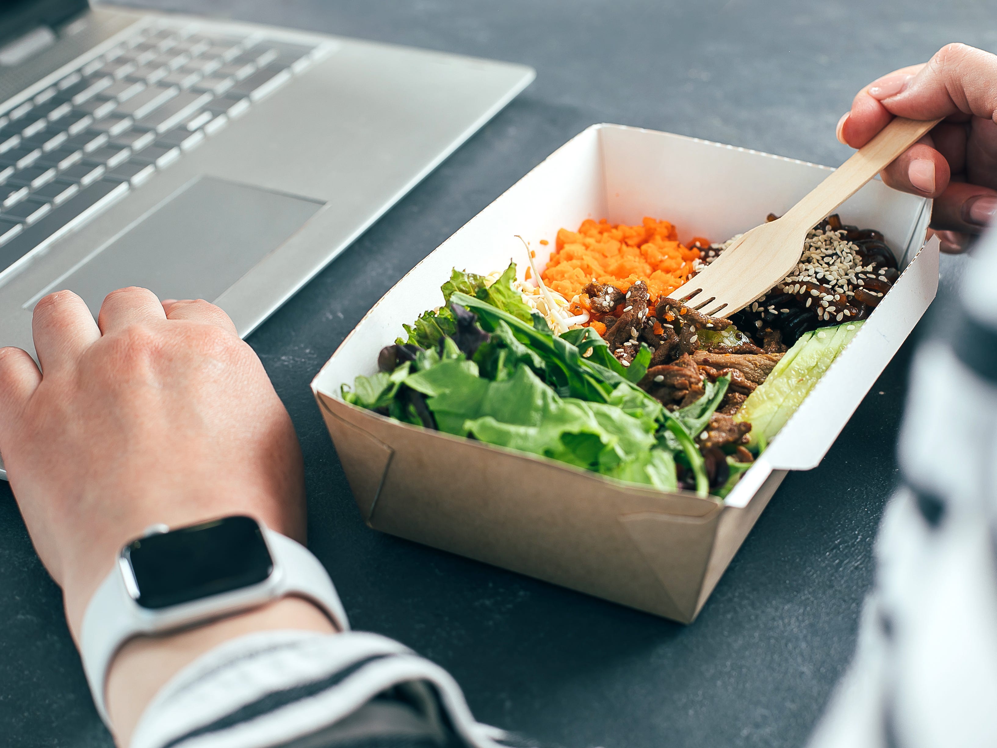 A close-up of a person eating from a cardboard meal box while working on a laptop.
