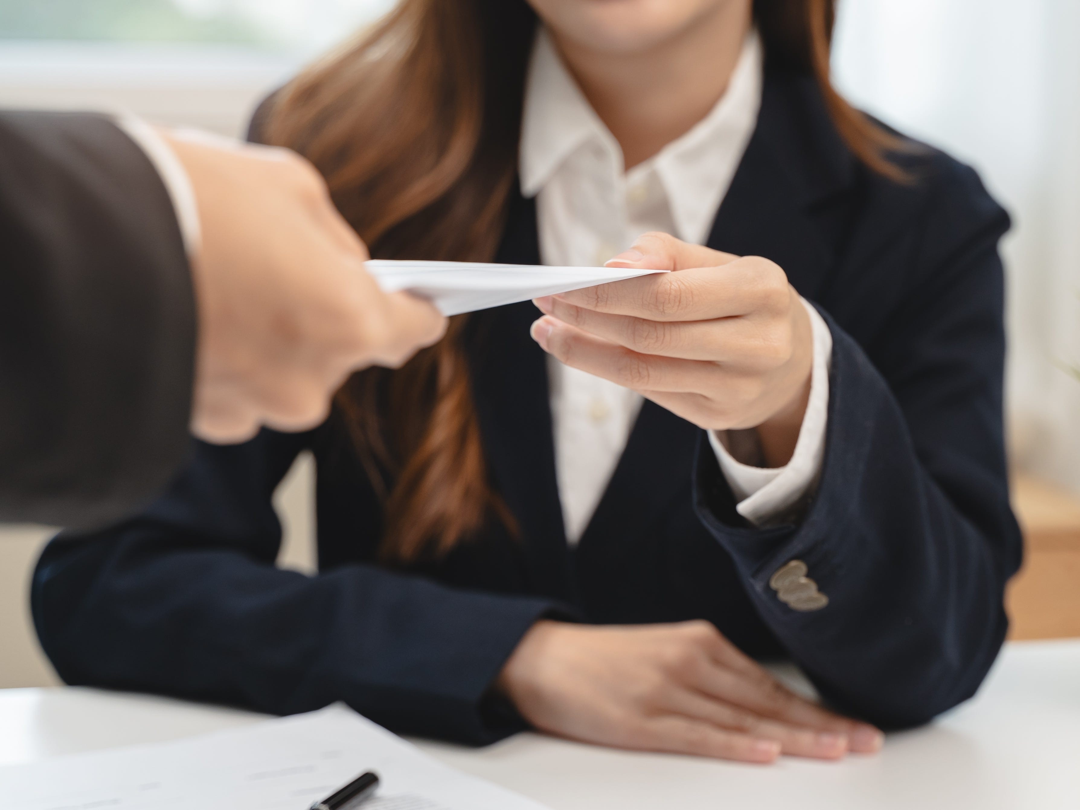 A person hands an envelope across a table to a woman wearing a suit.