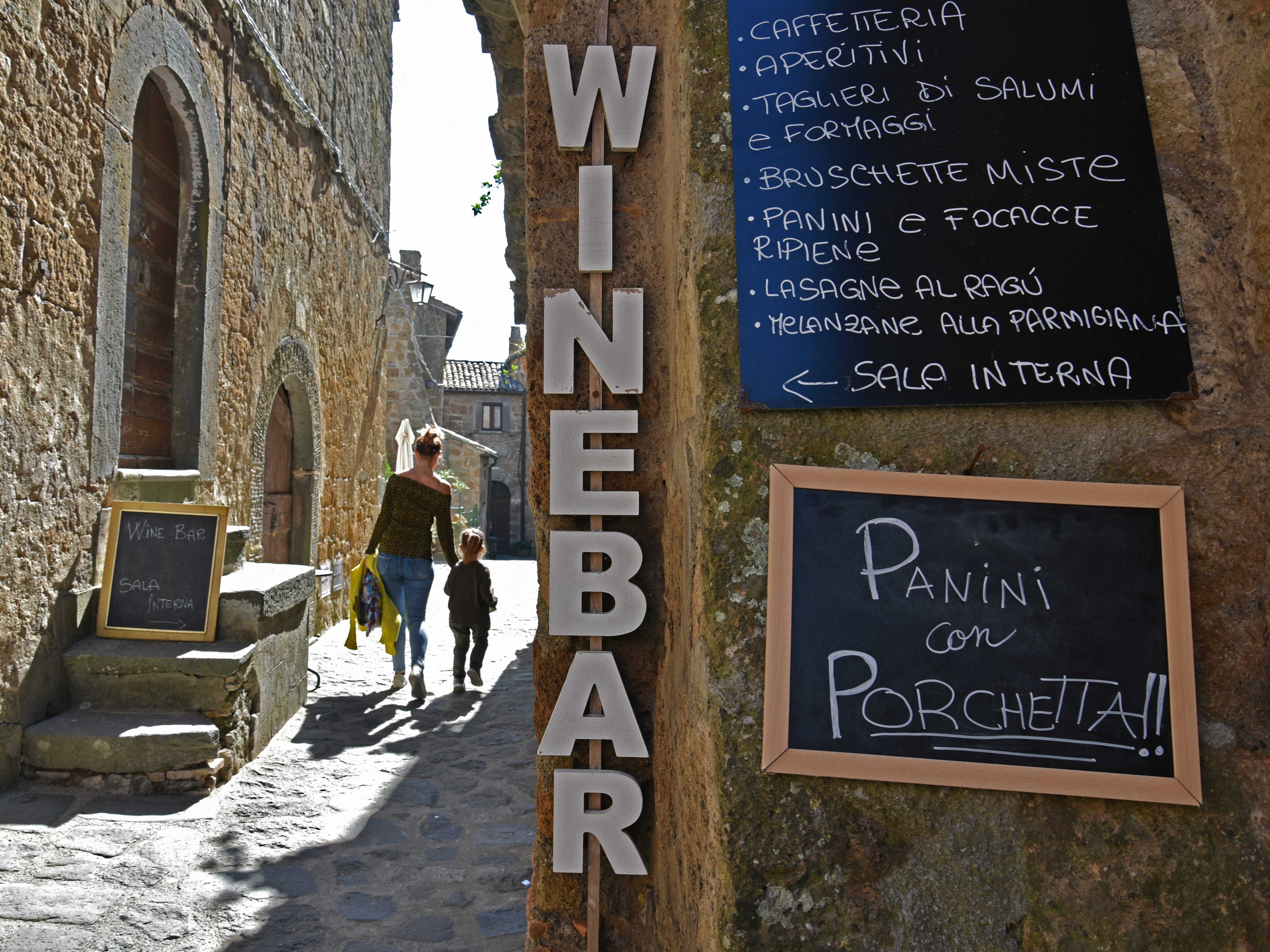 People walk down a cobblestone pathway next to a wine bar with a menu outside the building.
