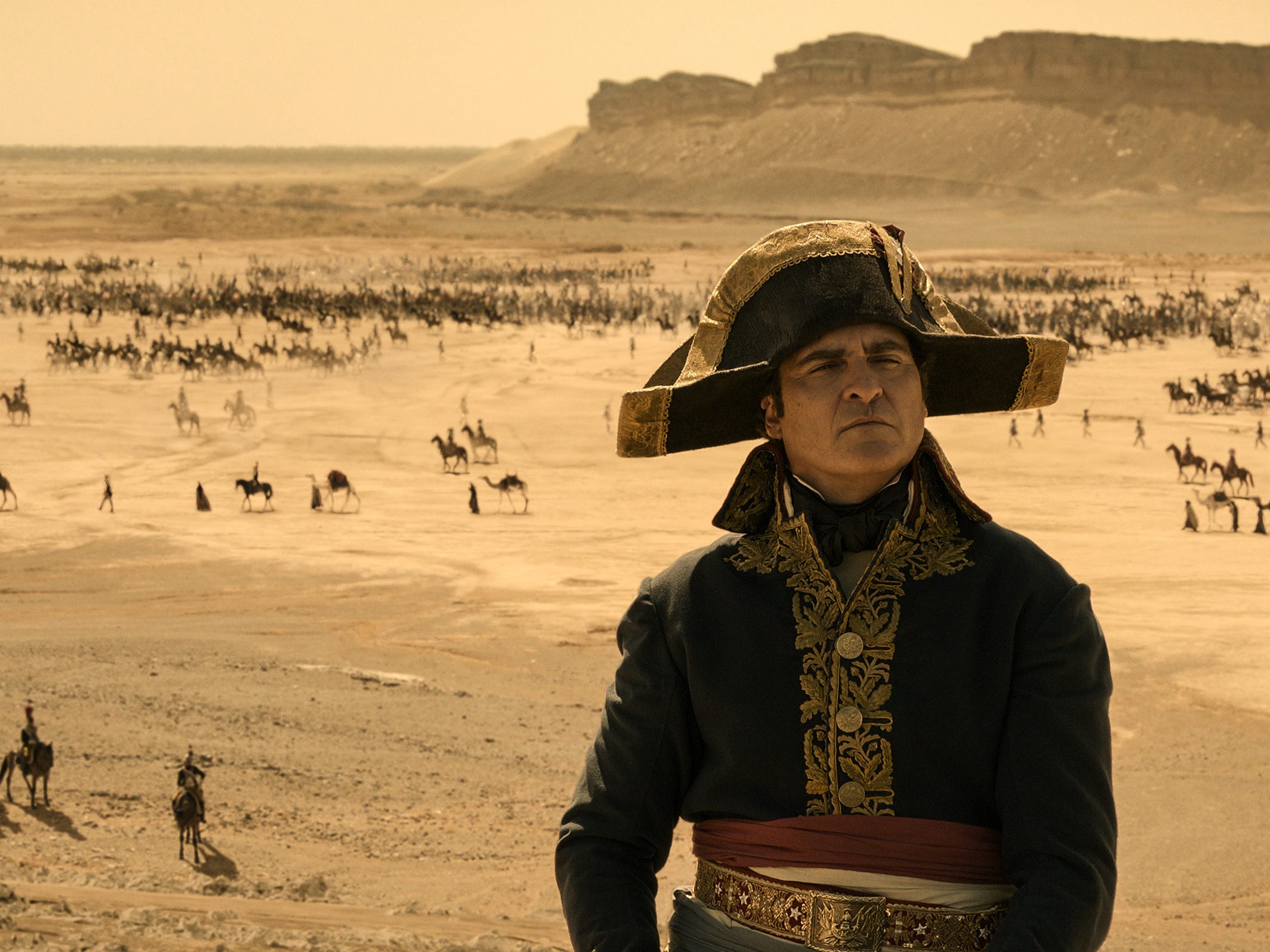 A still from "Napoleon" showing Joaquin Phoenix as Napoleon looking into the distance as troops move through the desert in the background.