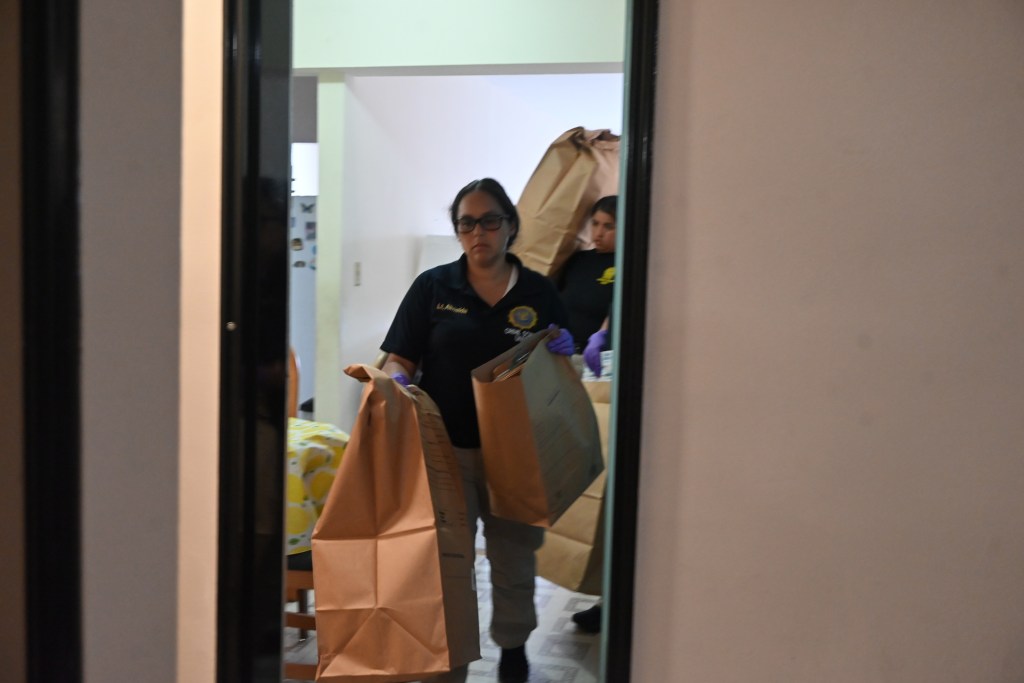 Two forensic investigators carrying evidence bags in an apartment.