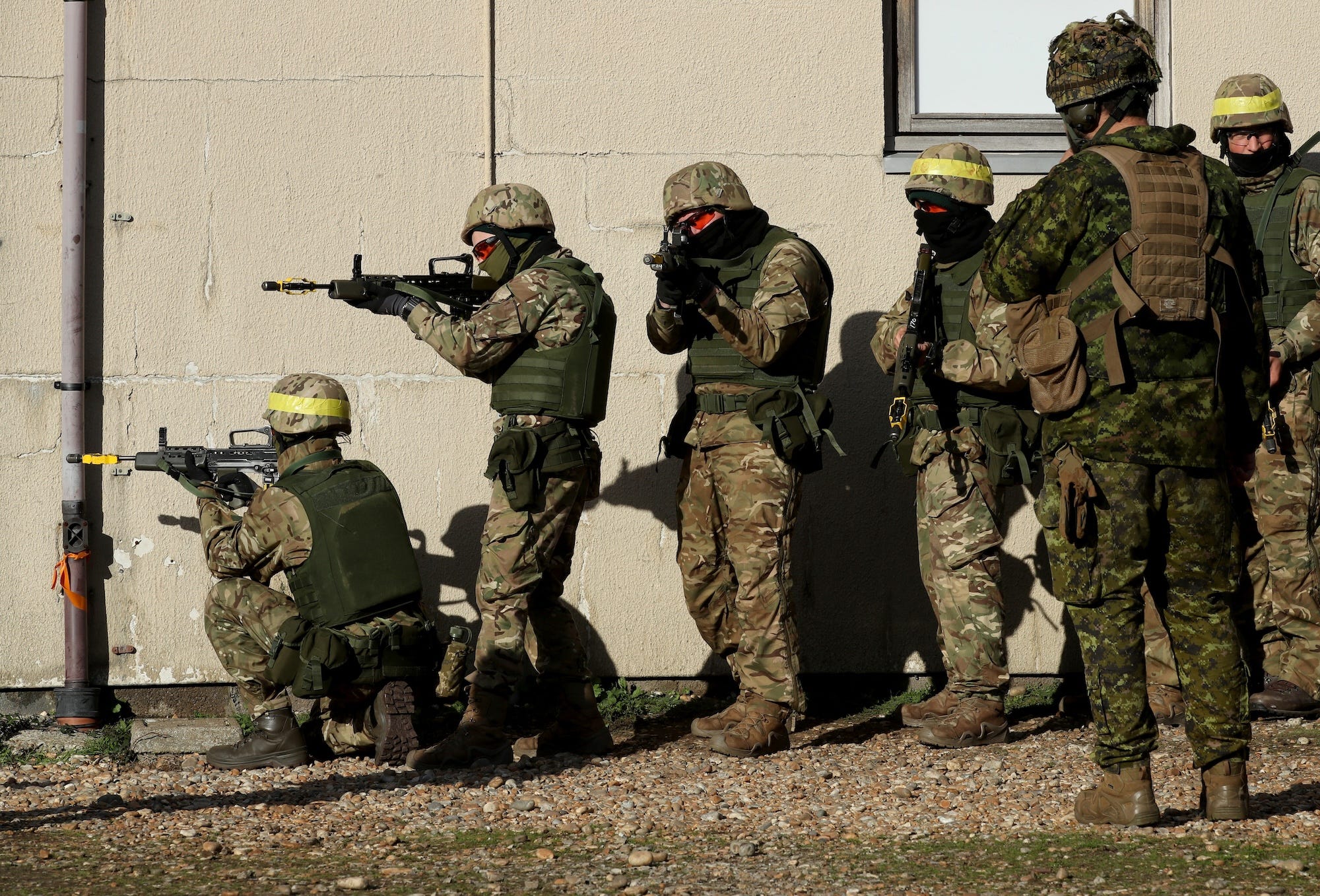 Men in camouflage gear and helmets and red sunglasses stand in front of a pale yellow brick wall, some of them with firearms raised