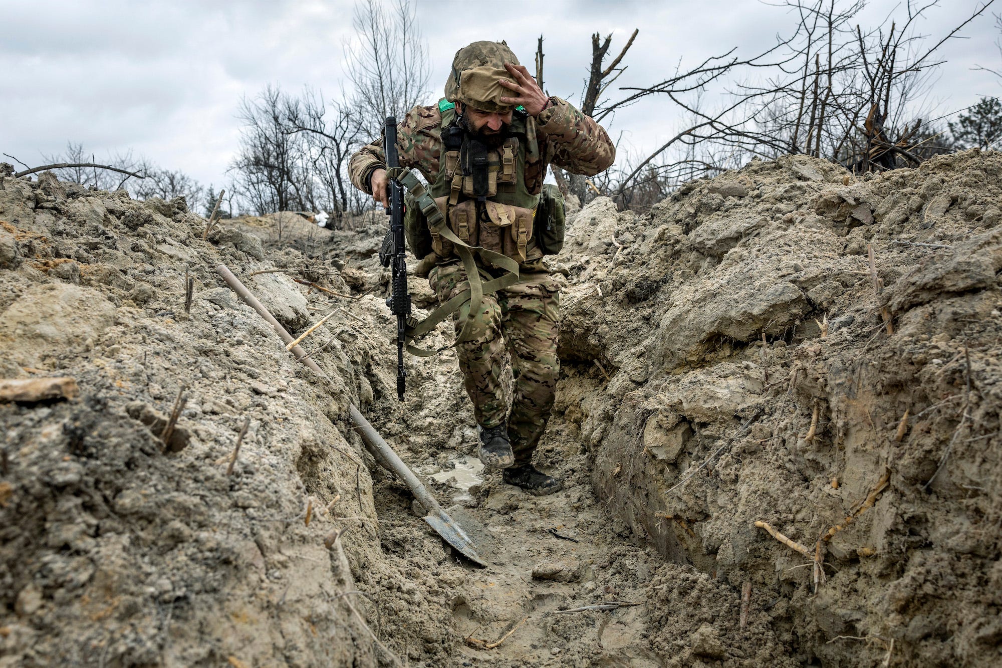 An armed Ukrainian medic running through a partially dug trench in front of a gray sky.