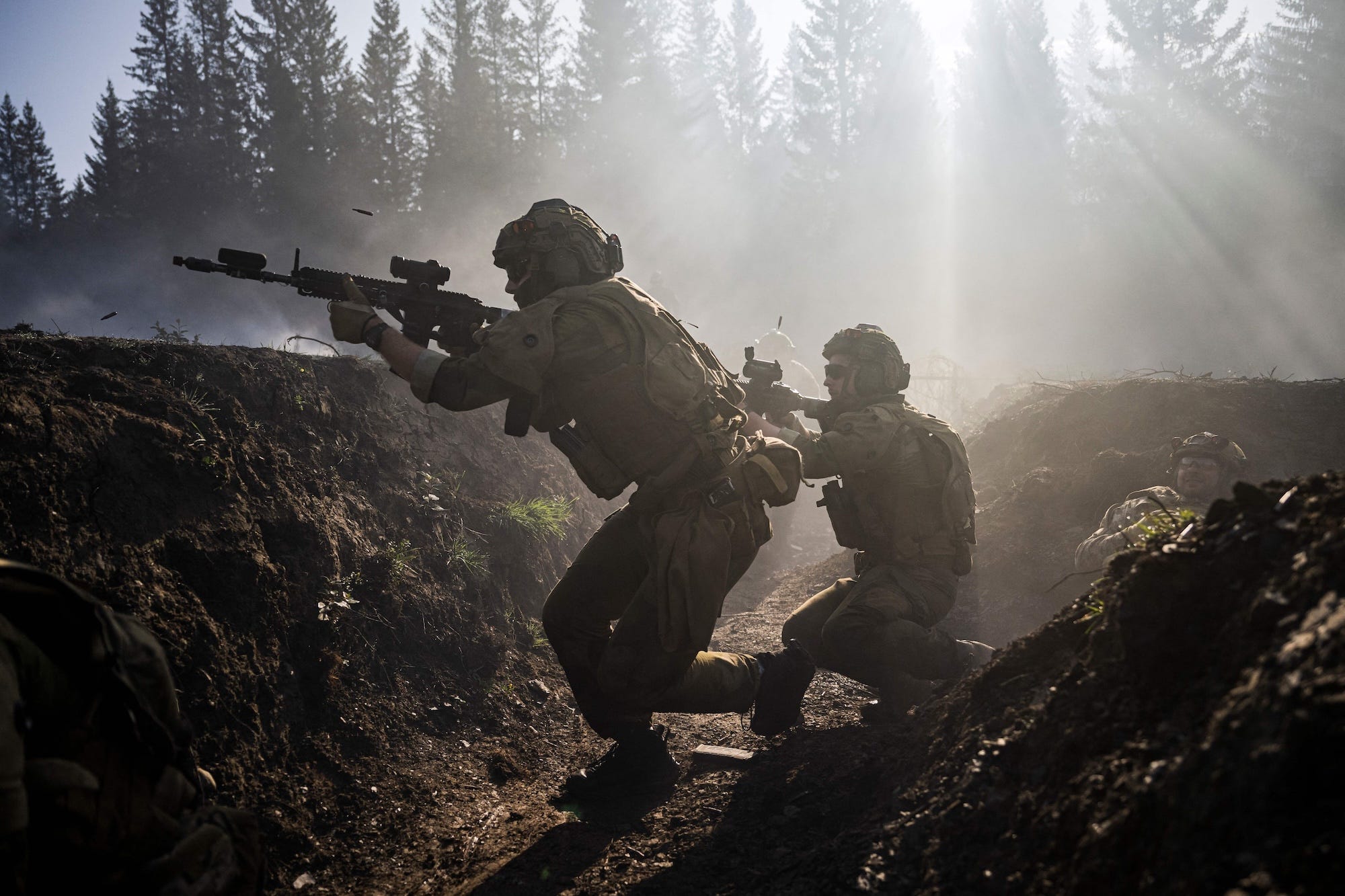 Two men in camouflage gear and helmets hold up firearms while in an earthern trench with misty trees behind them