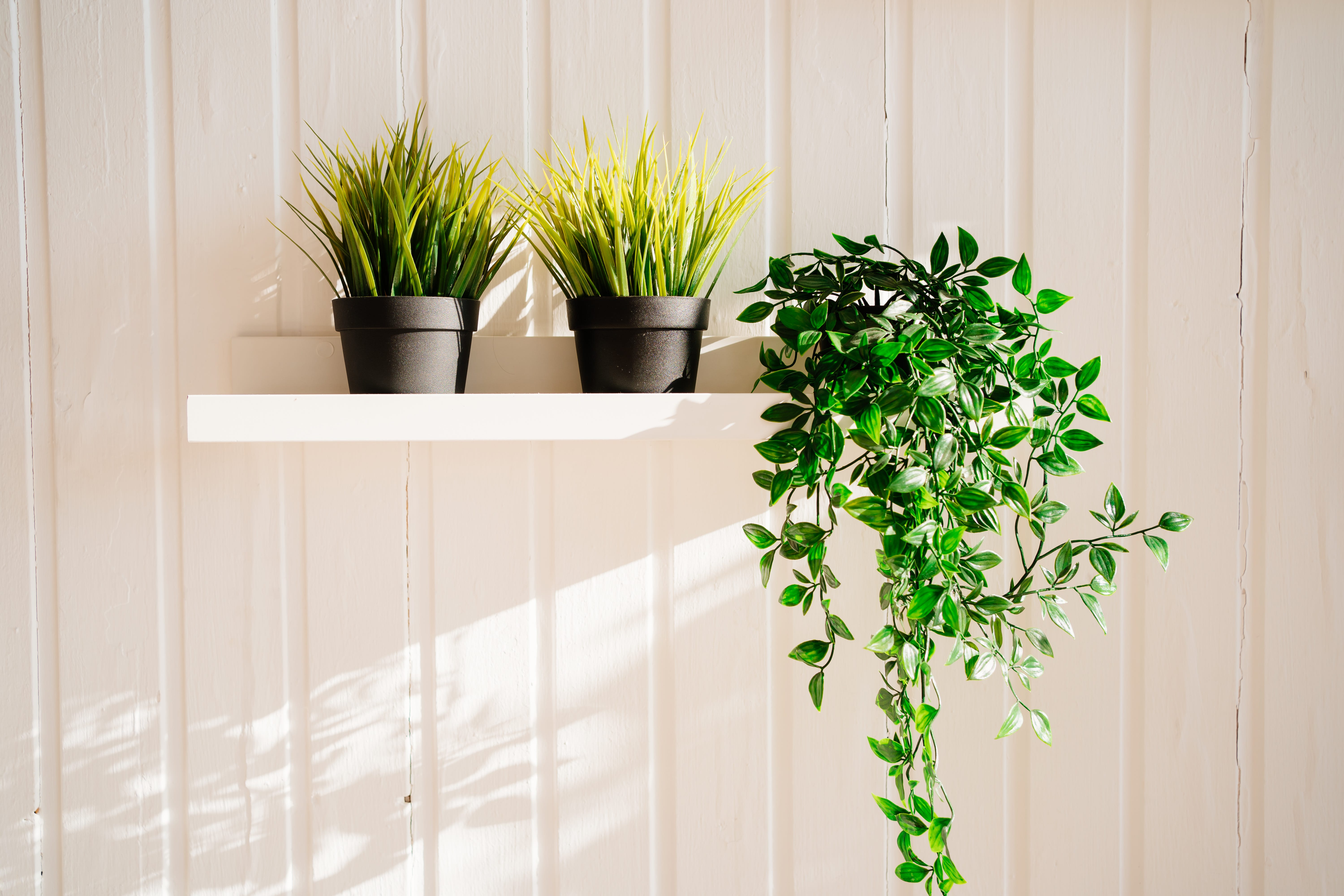 Three artificial green plants on a white shelf on a light pink wall.