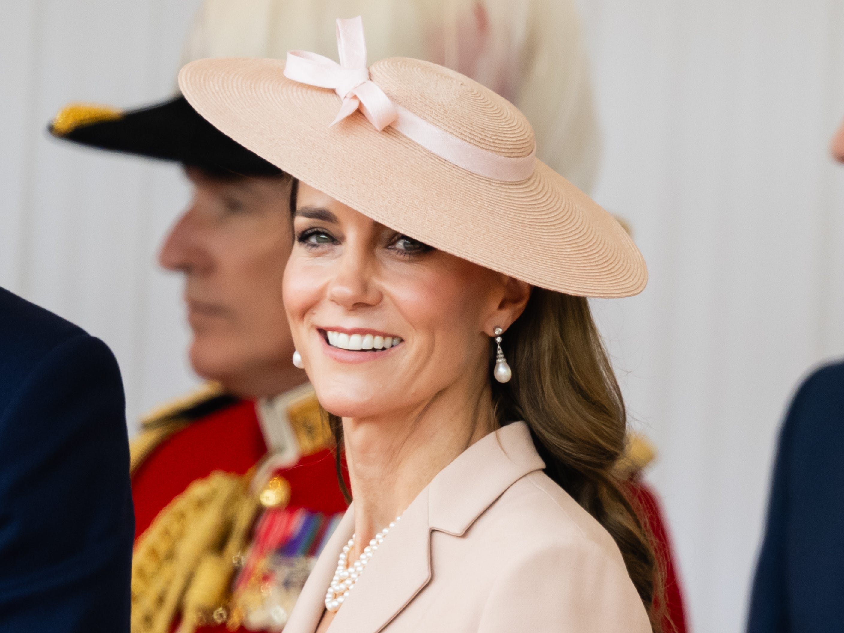 Kate Middleton smiles at a camera wearing a pink hat and jacket.