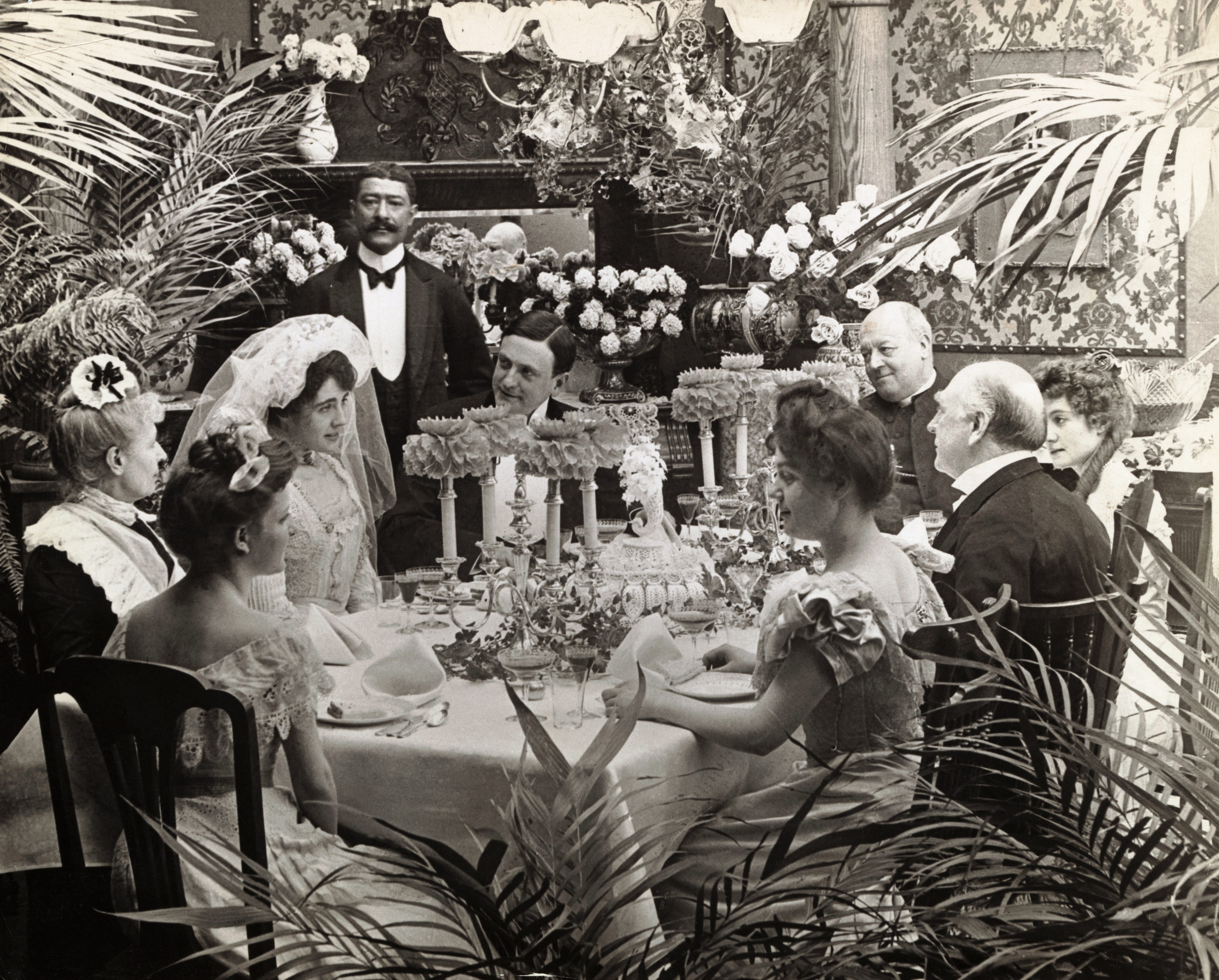 Undated photograph of a wedding reception. Well dressed people formally sitting around a banquet table.