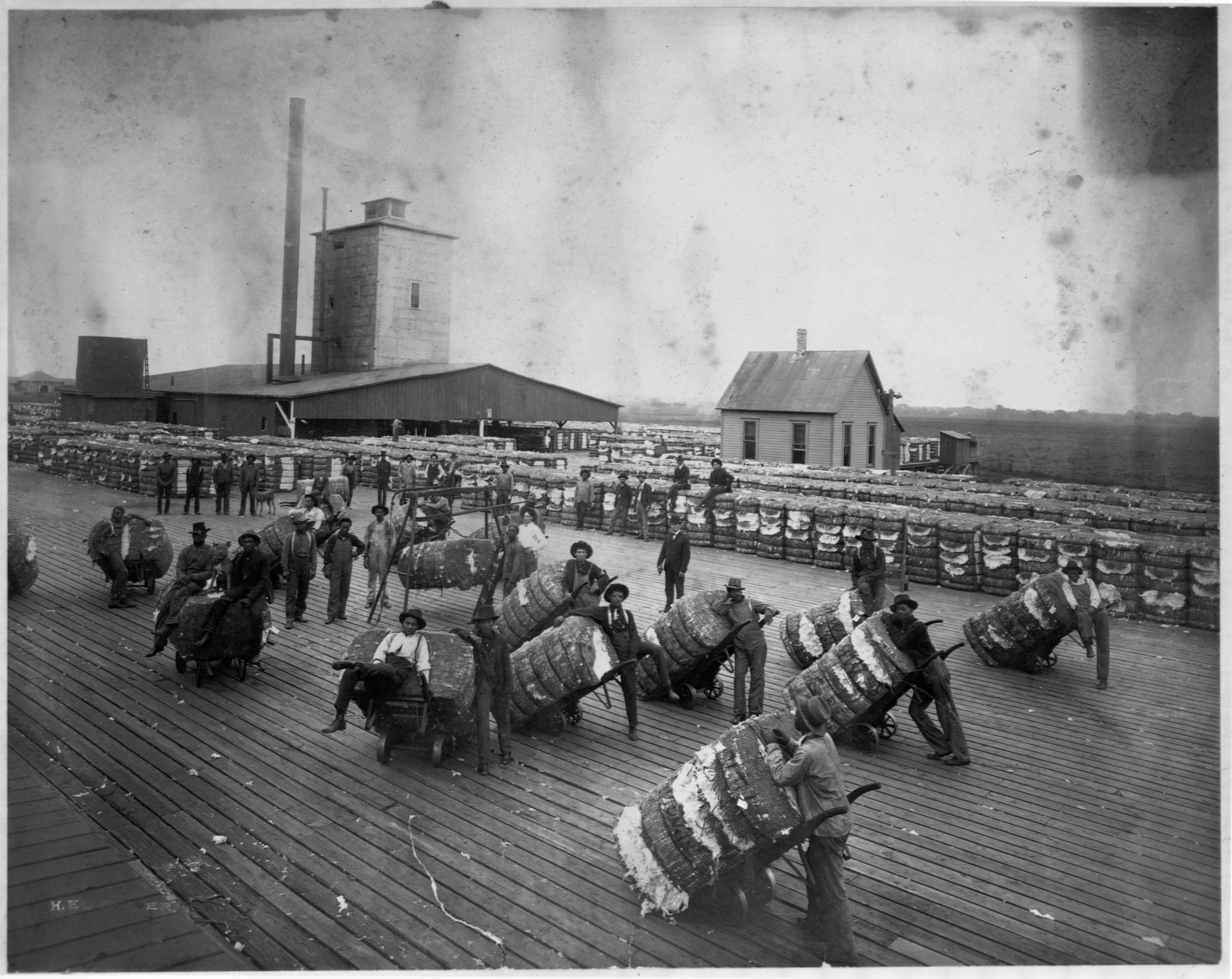 Black men stand with bales of cotton on a loading dock at a cotton compress.