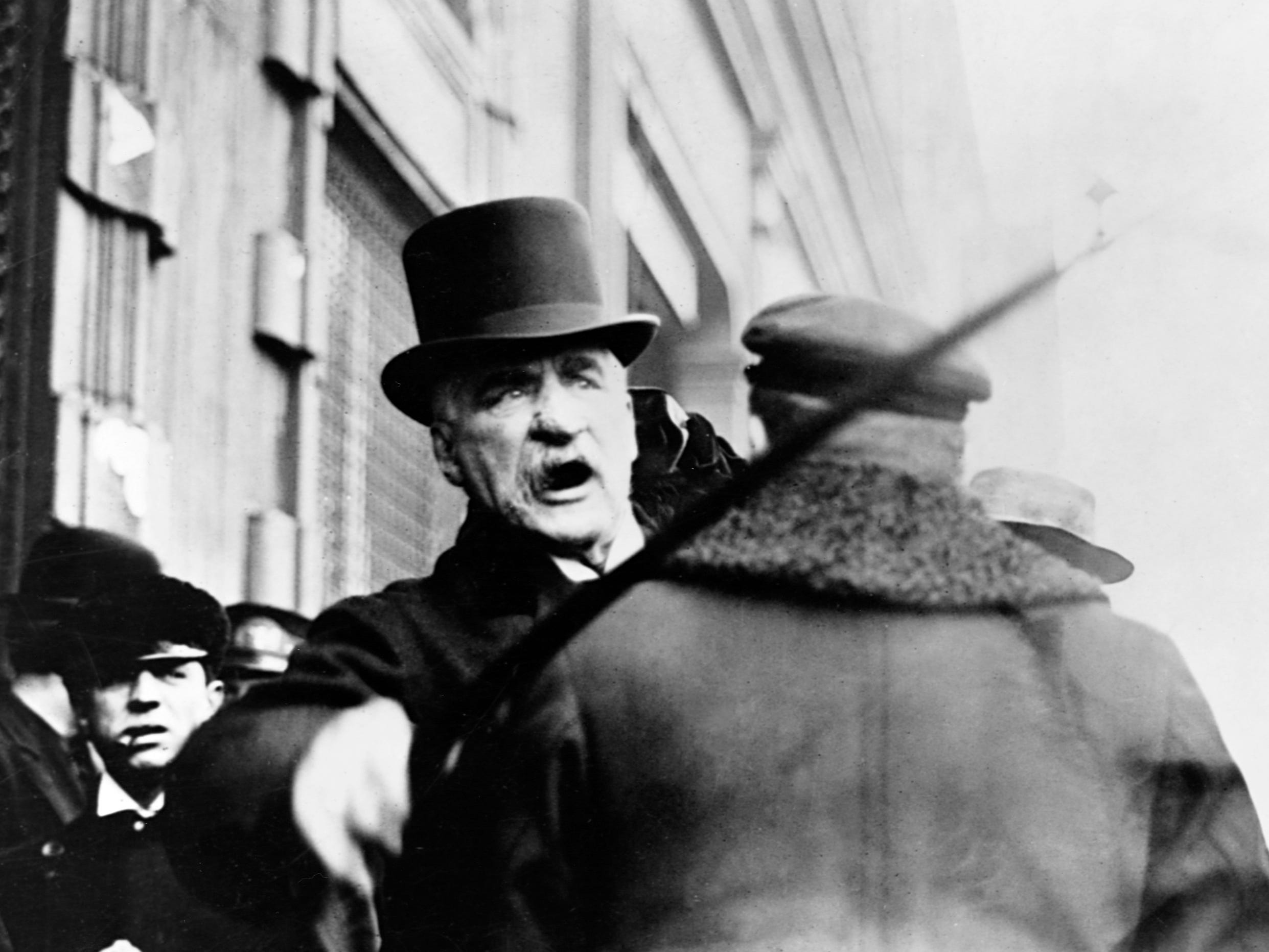 J.P. Morgan (1837-1913), founder of U.S. Steel, shakes his cane at someone as he walks down a city street.