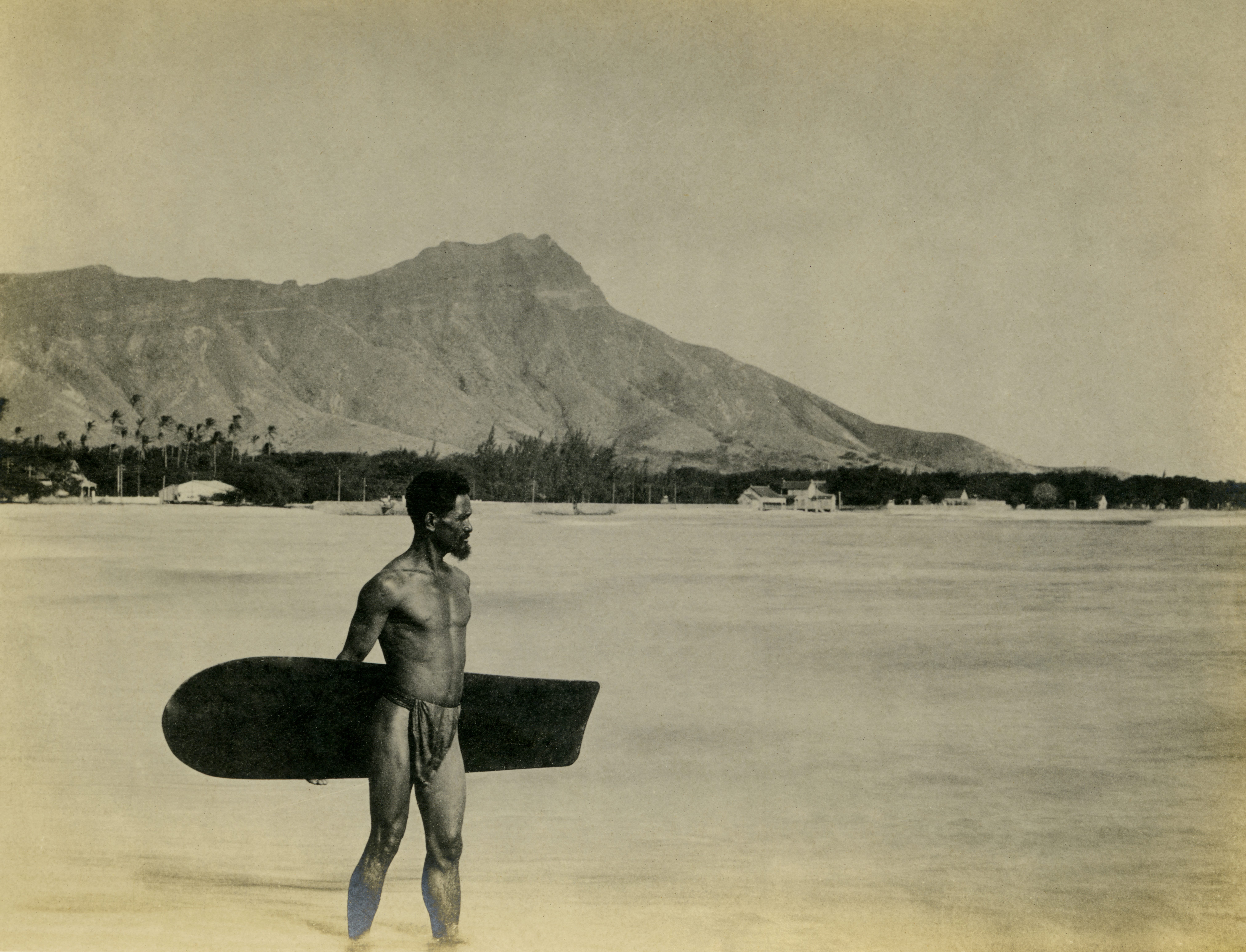 A black and white photograph shows a native Hawaiian surfer standing ankle deep in the ocean, holding his wooden surfboard behind his back, with Diamond Head in the background, circa 1900.