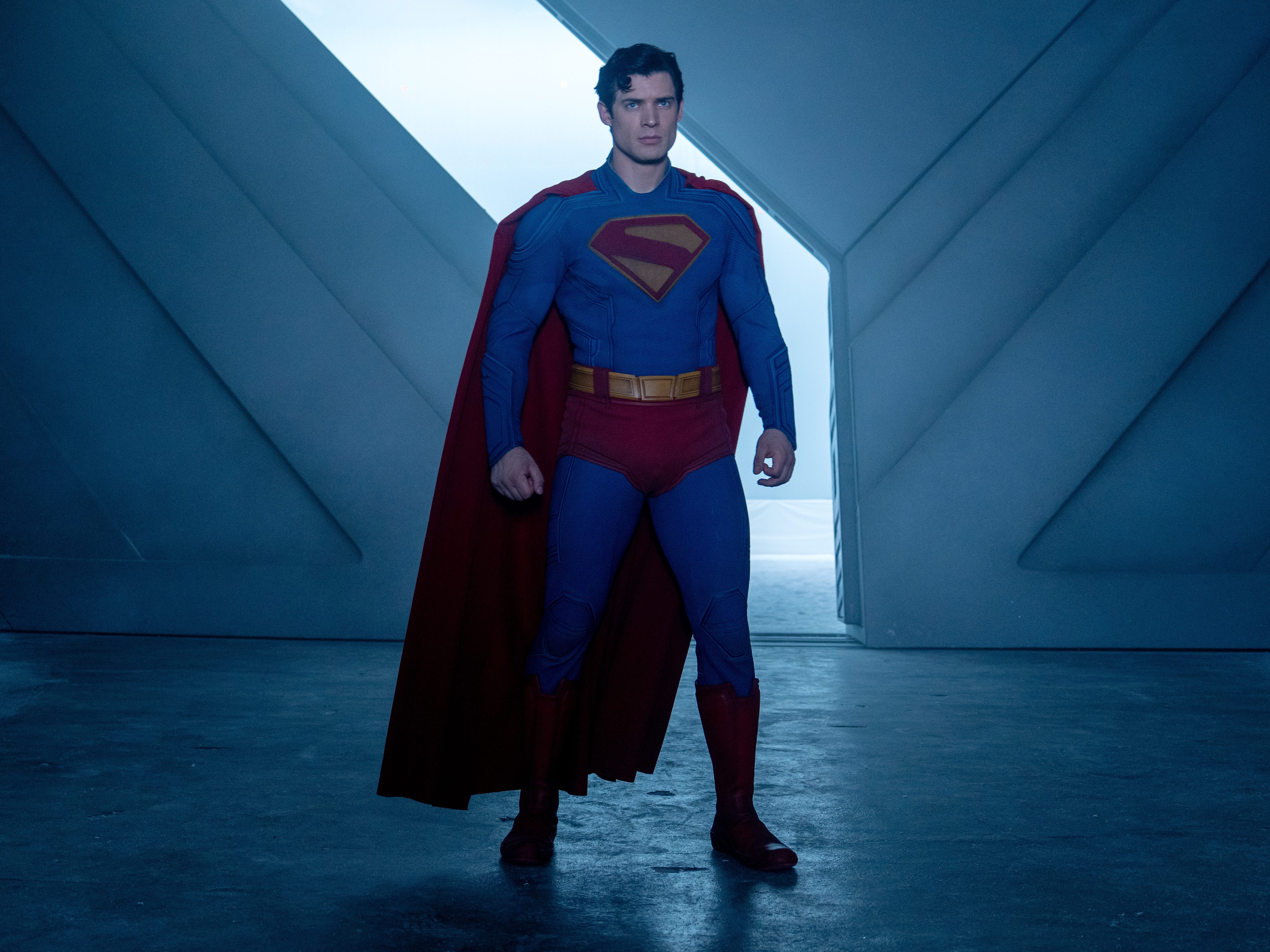 A man with black hair stands in an icy room with a large metal door behind him. He's wearing a blue costume with red trunks, a red cape, red boots and a red S on his chest.