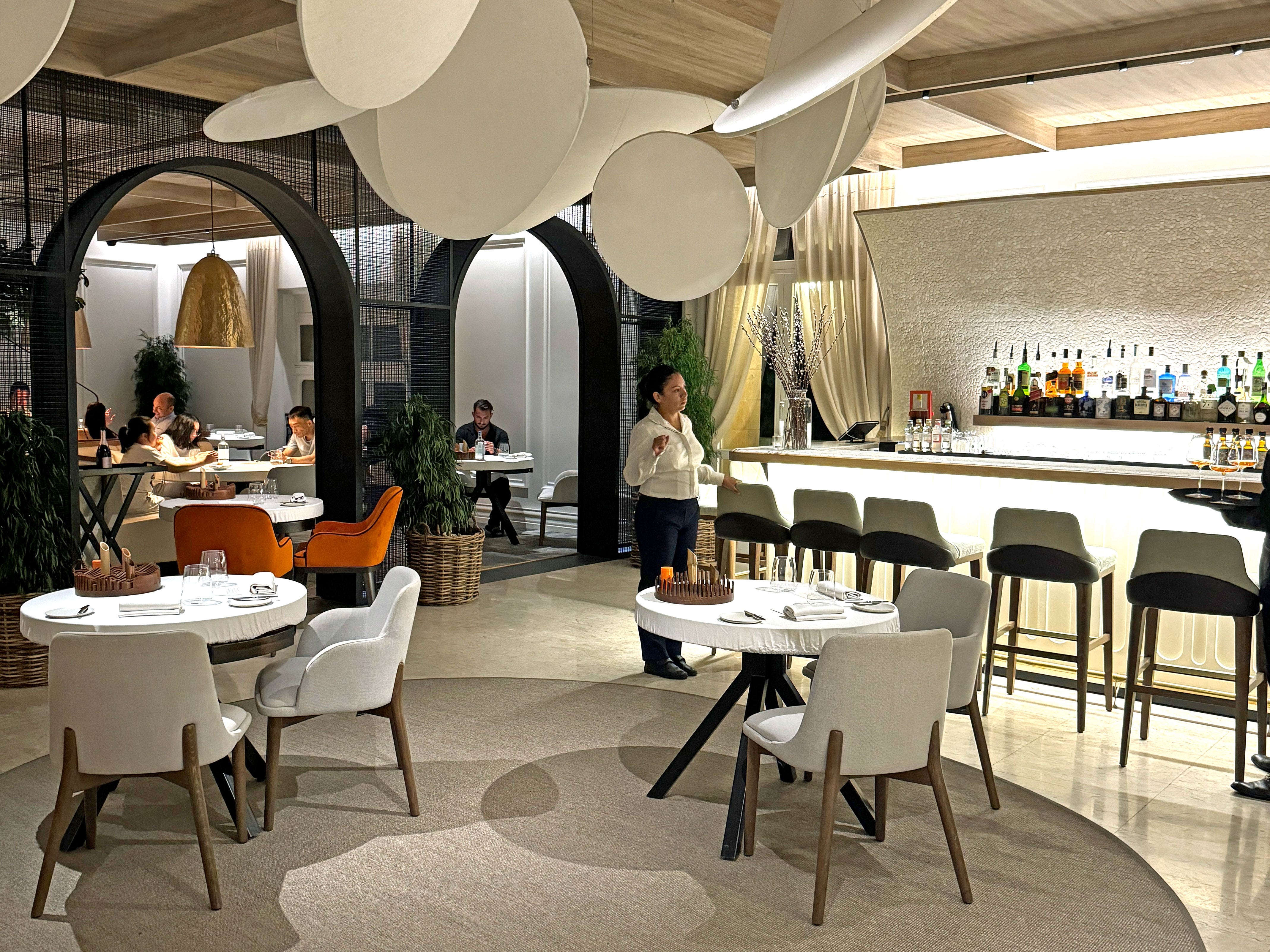 A server walks through a chic restaurant with round tables and a bar.