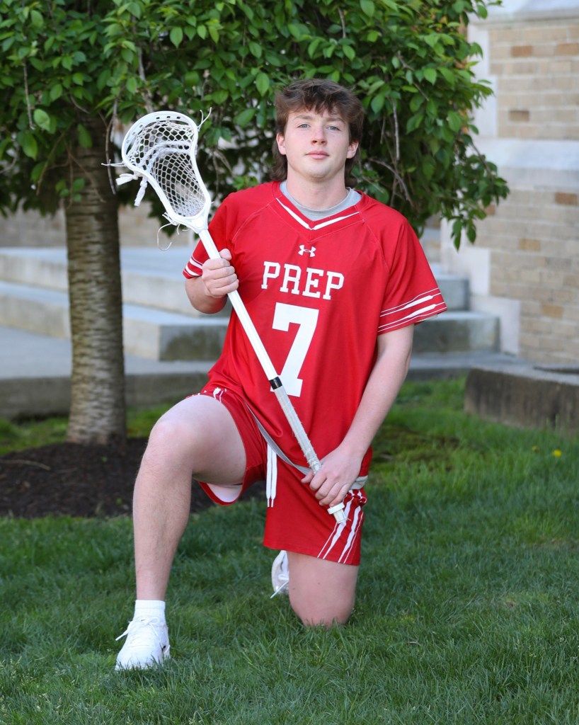 Lacrosse player kneeling on grass, holding lacrosse stick.