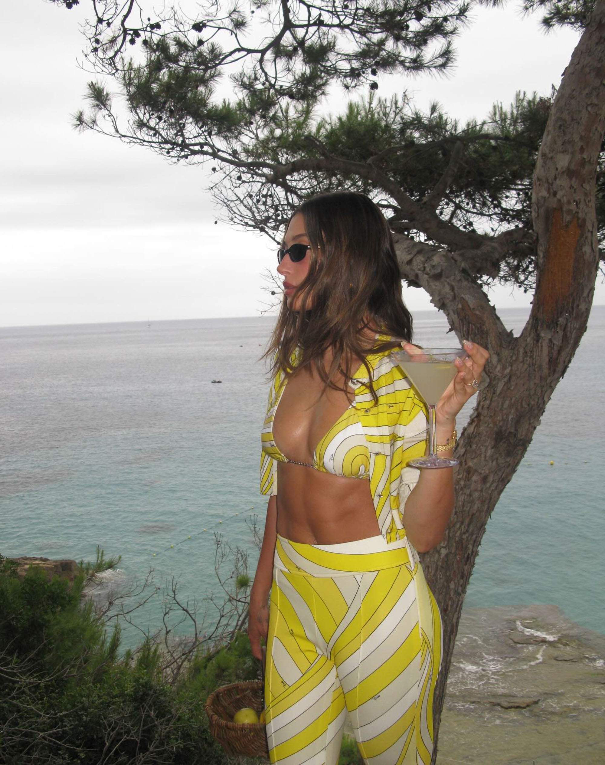 Hailey Bieber holds a martini while standing near the ocean. 