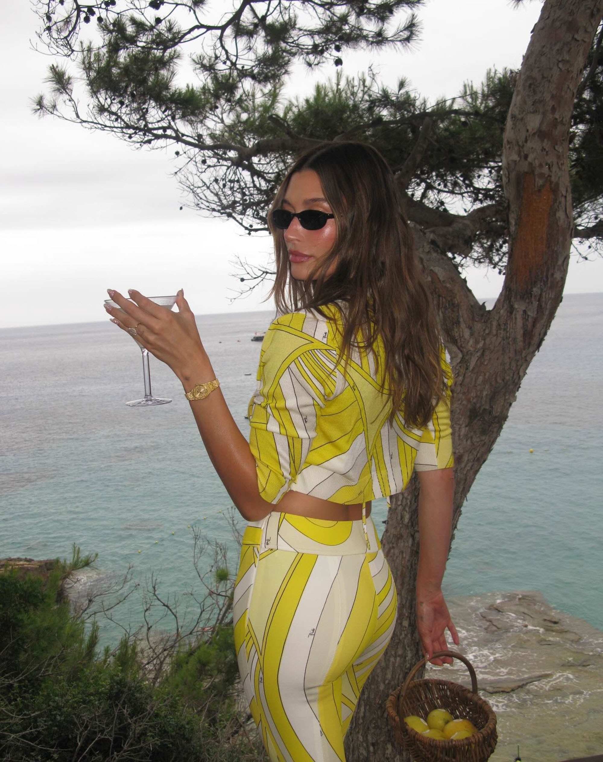 Hailey Bieber holds a basket of lemons near the water. 