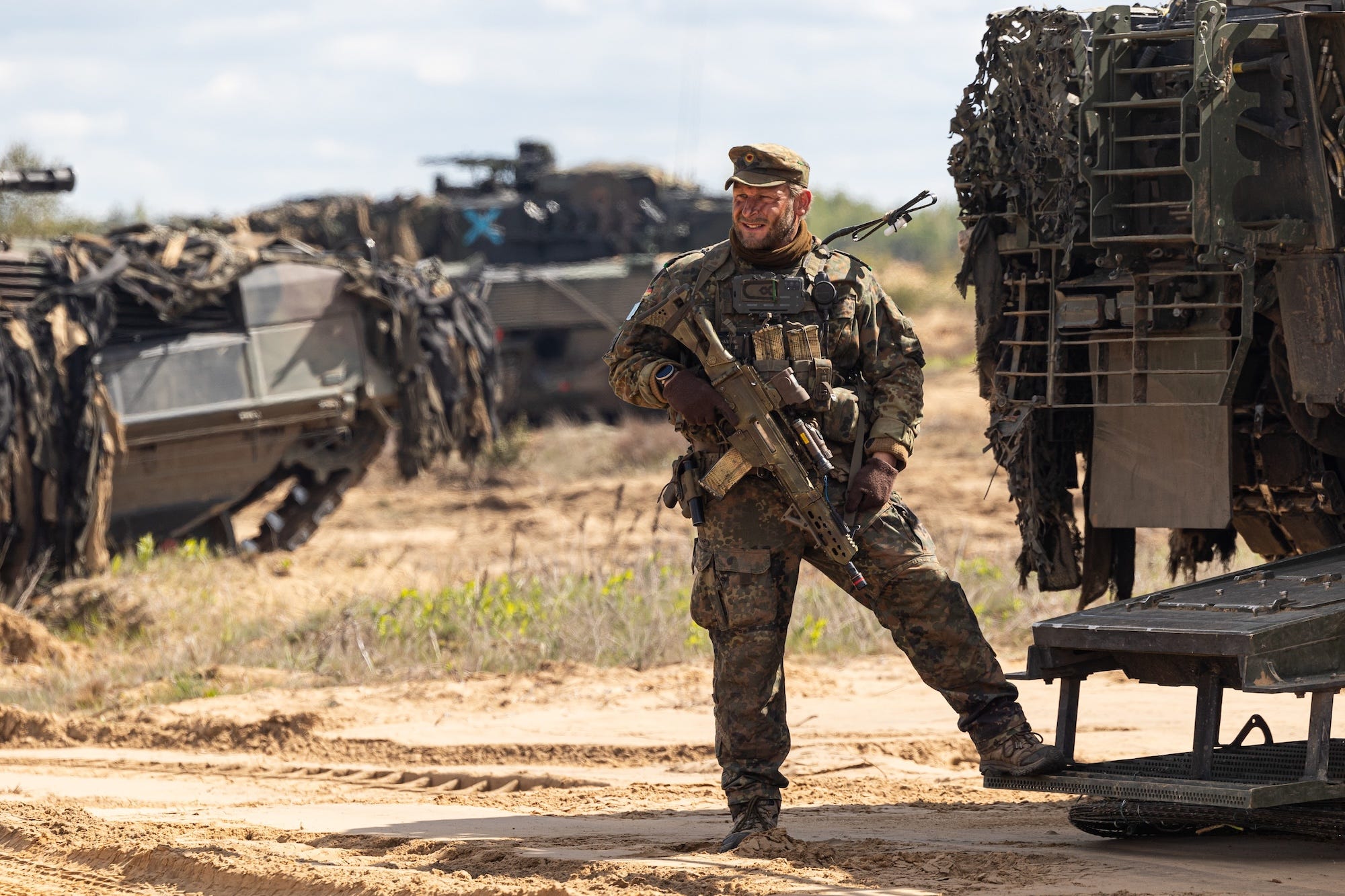 A man wearing camouflage gear and holding a firearm stands with his leg on a step on a camouflage-covered armored vehicle with more similar vehicles behind him