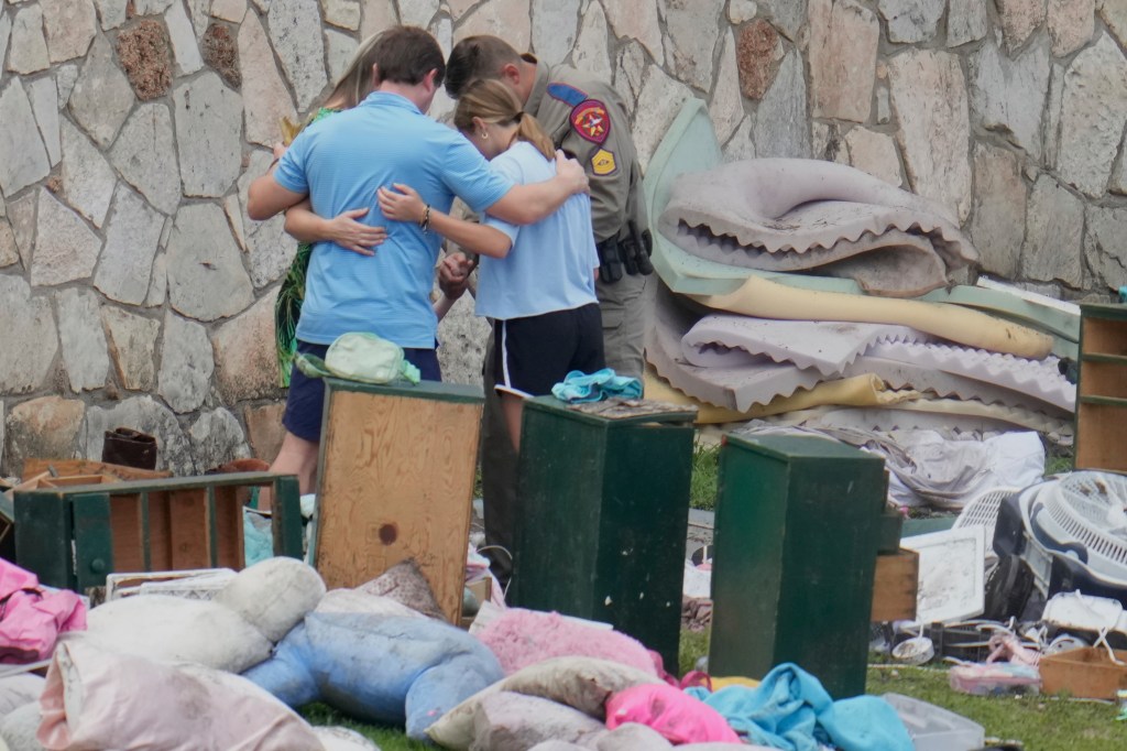 Family embraces while an officer looks on amidst flood debris.