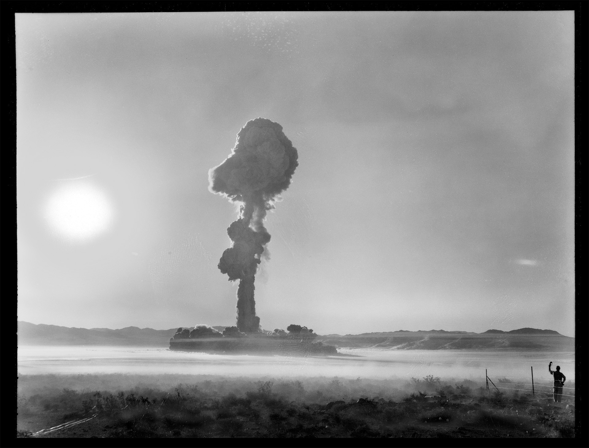 black-and-white photo of daytime desert landscape with hazy sun near dark mushroom cloud in distance, with a man standing in foreground watching with hand shielding eyes