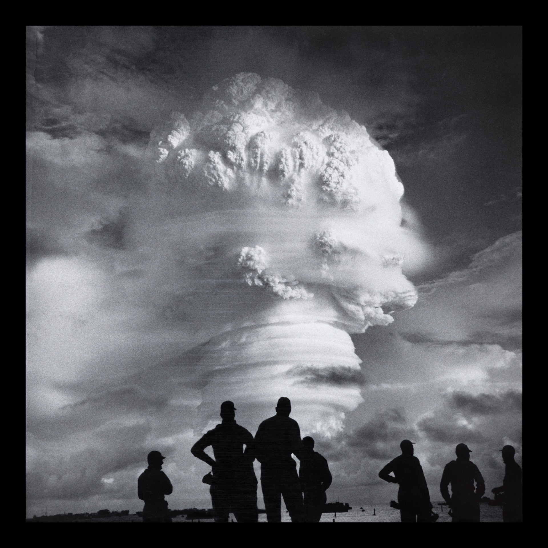 black-and-white photo of silhouetted group of soldiers standing on shore with enormous swirling mushroom cloud in distance