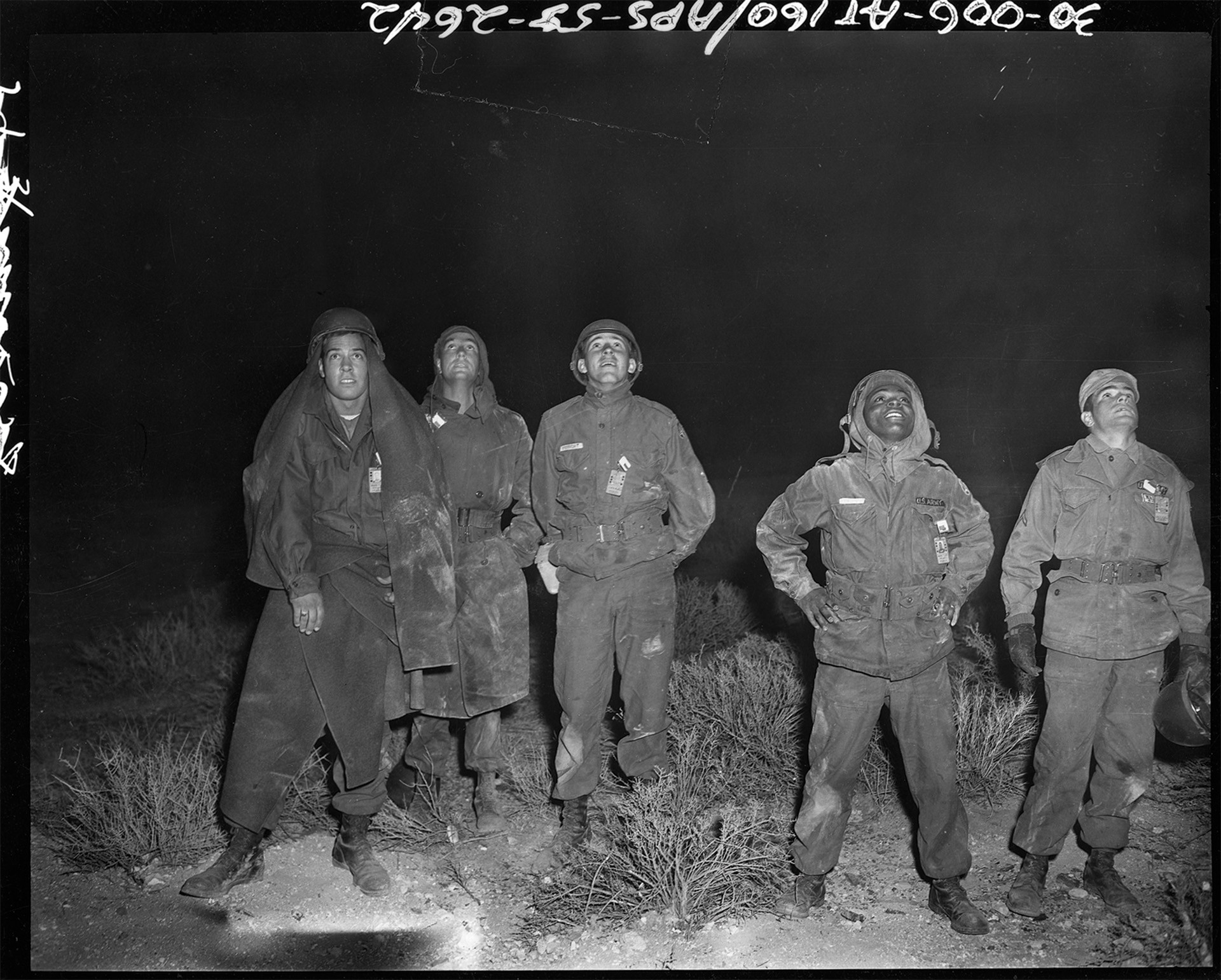 black-and-white nighttime photo of 5 uniformed soldiers standing and looking skyward, brightly lit by the explosion