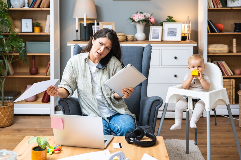 A mother works from home on her laptop, tablet, and phone while caring for her baby.