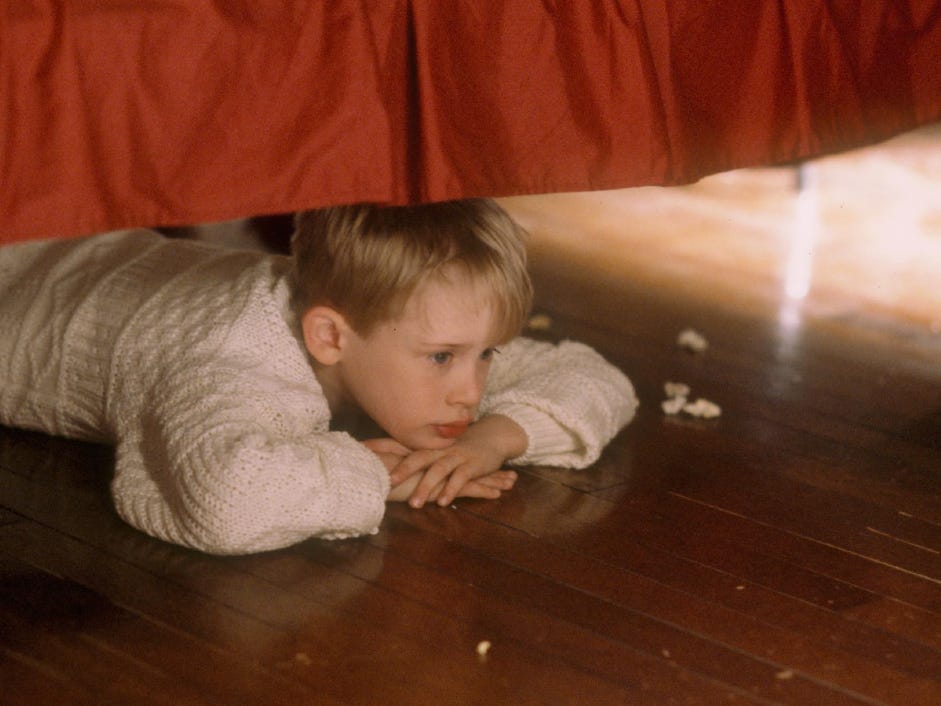 Kevin McCallister wears a white cable-knit sweater and lies on his stomach under a bed with a red bedskirt in a still from the original "Home Alone" movie.