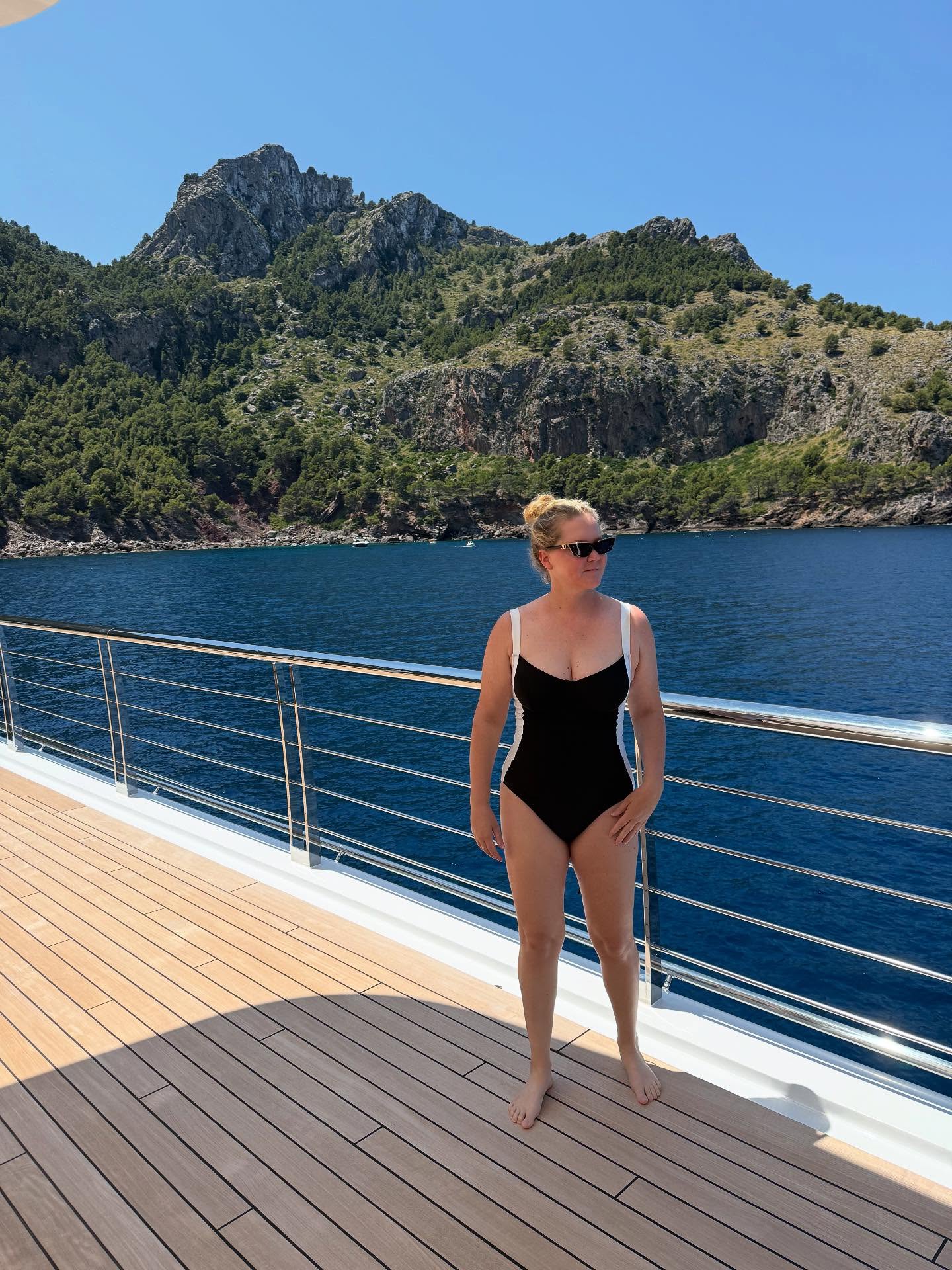 Amy Schumer stands on a boat deck in a black swimsuit. 