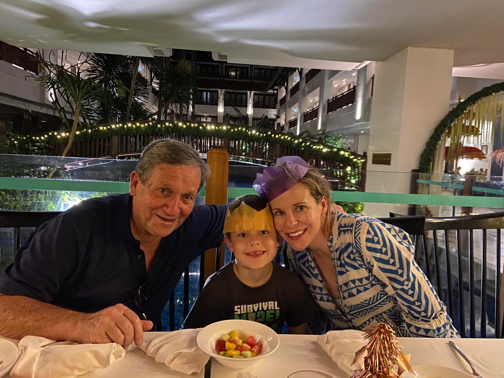 The author at dinner at night with her father and son, smiling at the camera. There are lights behind them and a bowl of food on the table.