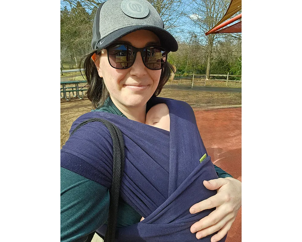A selfie of a woman with brown hair wearing a baseball cap, sunglasses, and a sleeping baby in a carrier wrapped around her chest.