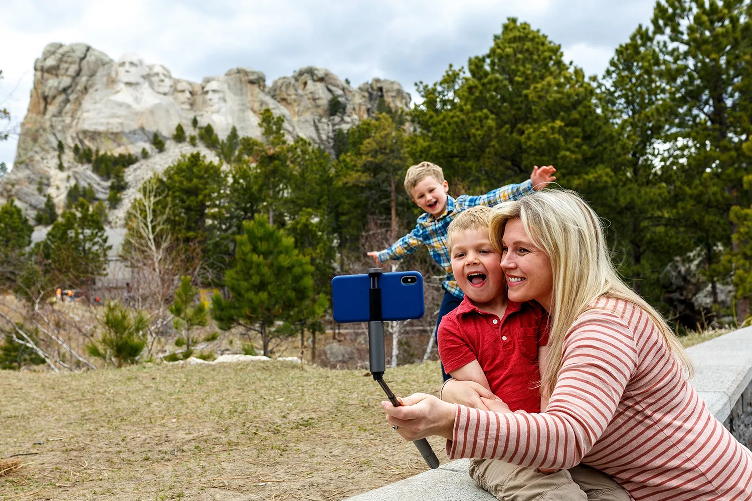 A blond woman wearing a striped shirt uses a selfie stick to take a photo with her camera. She holds a young child in a red polo shirt. Another child in a plaid shirt stands behind them, arms spread wide as he smiles. The Mount Rushmore memorial looms in the distance, with trees scattered between it and the family.