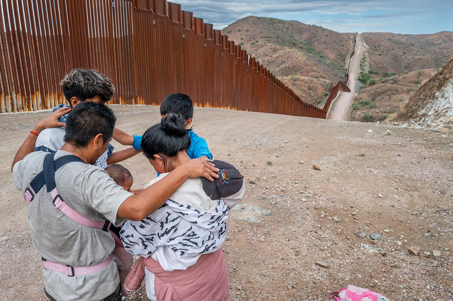A migrant family of five, with a man, woman, and middle-grade boys plus a baby in a carrier, put their arms on each other's shoulders as they stand in a circle with heads bowed. Behind them, the tall rusty metal barriers of a border fence extend along a rolling road up onto a dusty hillside on the horizon.