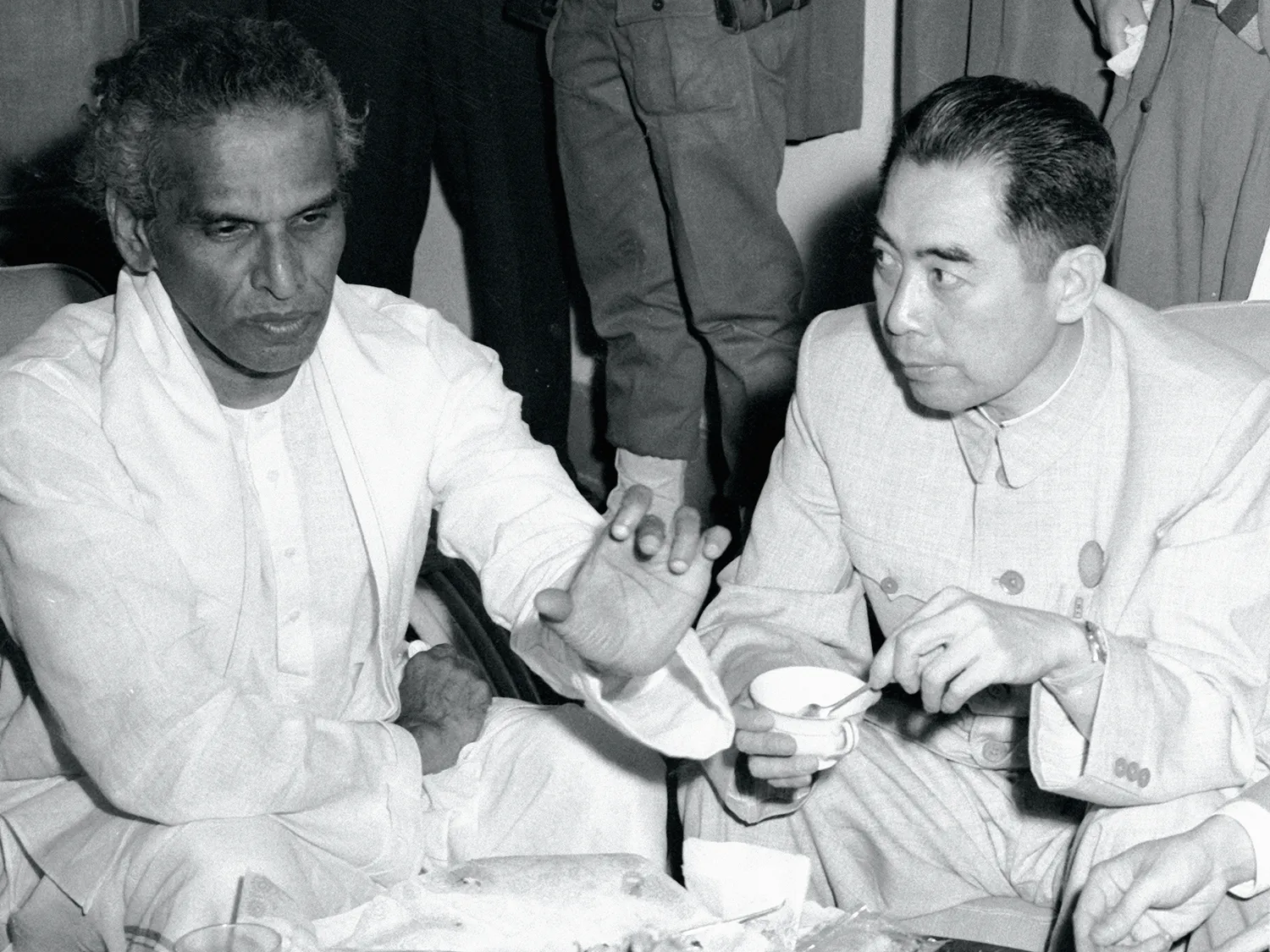 In this black-and-white photo, Menon gestures as Zhou drinks from a small cup and leans close to listen. Both men sit cross-legged on the ground with the tea set between them.