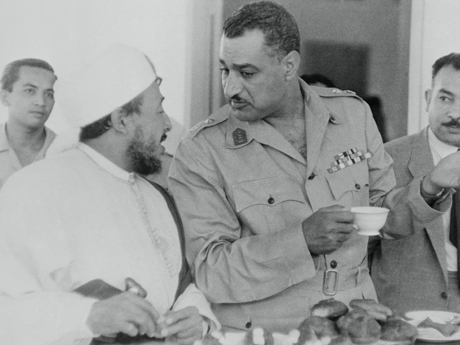 Hassen, wearing white and a hat, leans close as he confers with a taller Nasser, who wears a military uniform and holds a teacup.