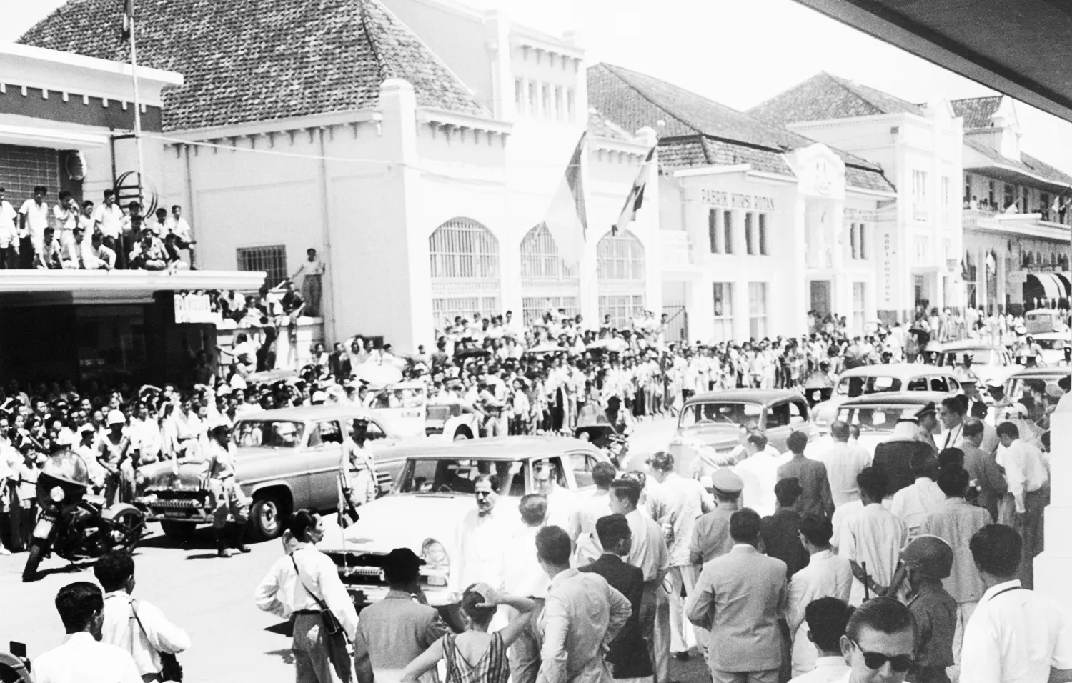 In this black-and-white historic photo, crowds of hundreds of people, mostly men, throng around cars as they move down a street. A large building is seen in the background, with more people watching from a street-facing balcony.