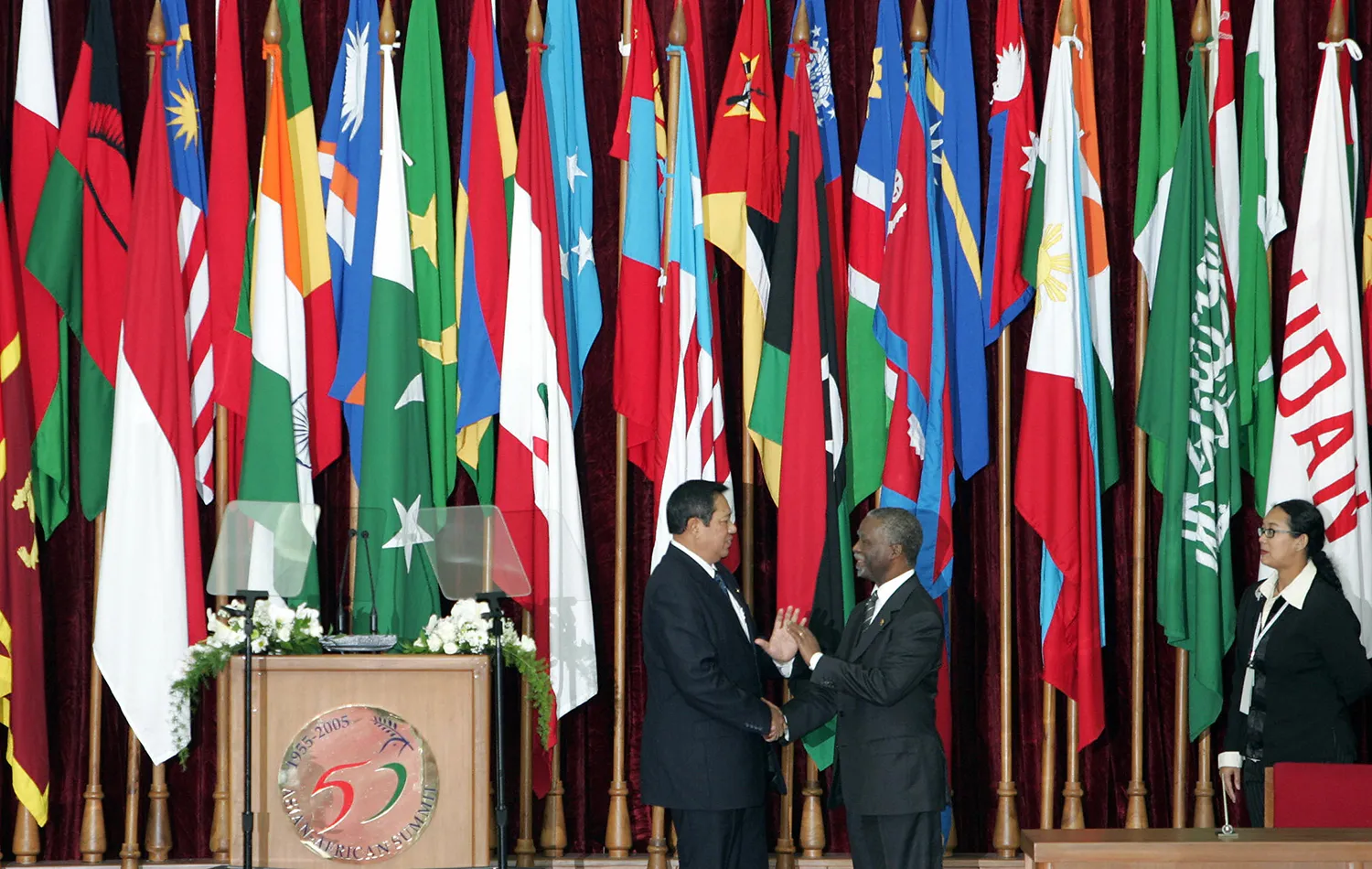 The two men shake hands onstage next to a lectern in front of a row of tall multi-colored flags on stands. Both men wear black suits and ties.
