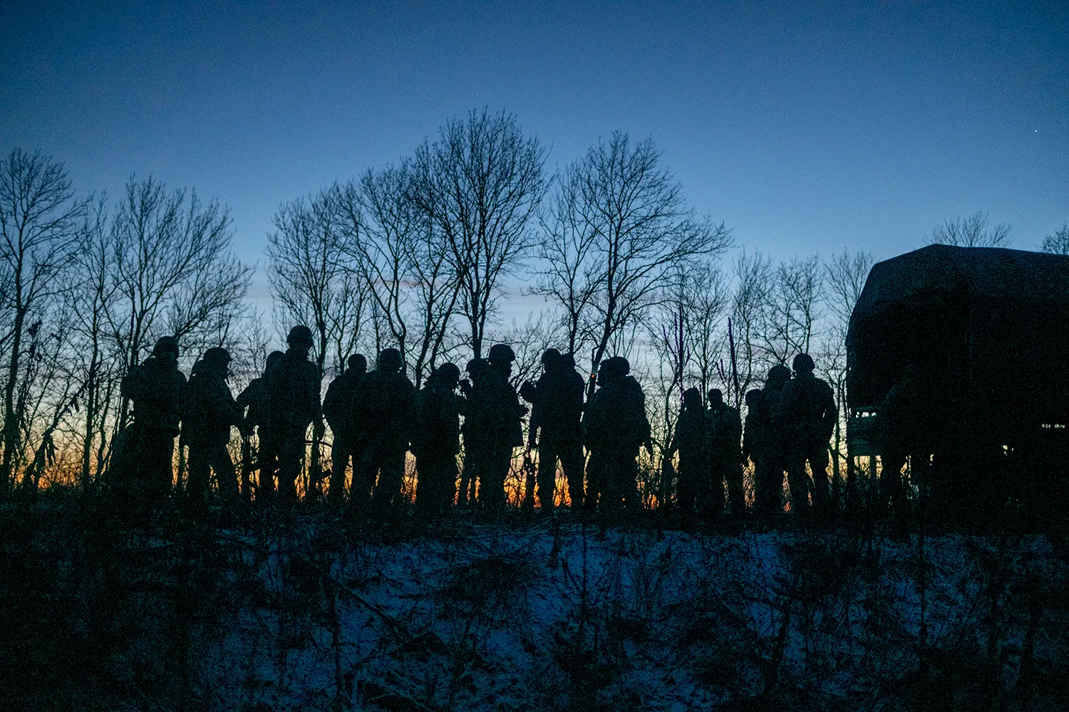 Soldiers in helmets are silhouetted against bare trees as they stand on a snowy field at dusk.