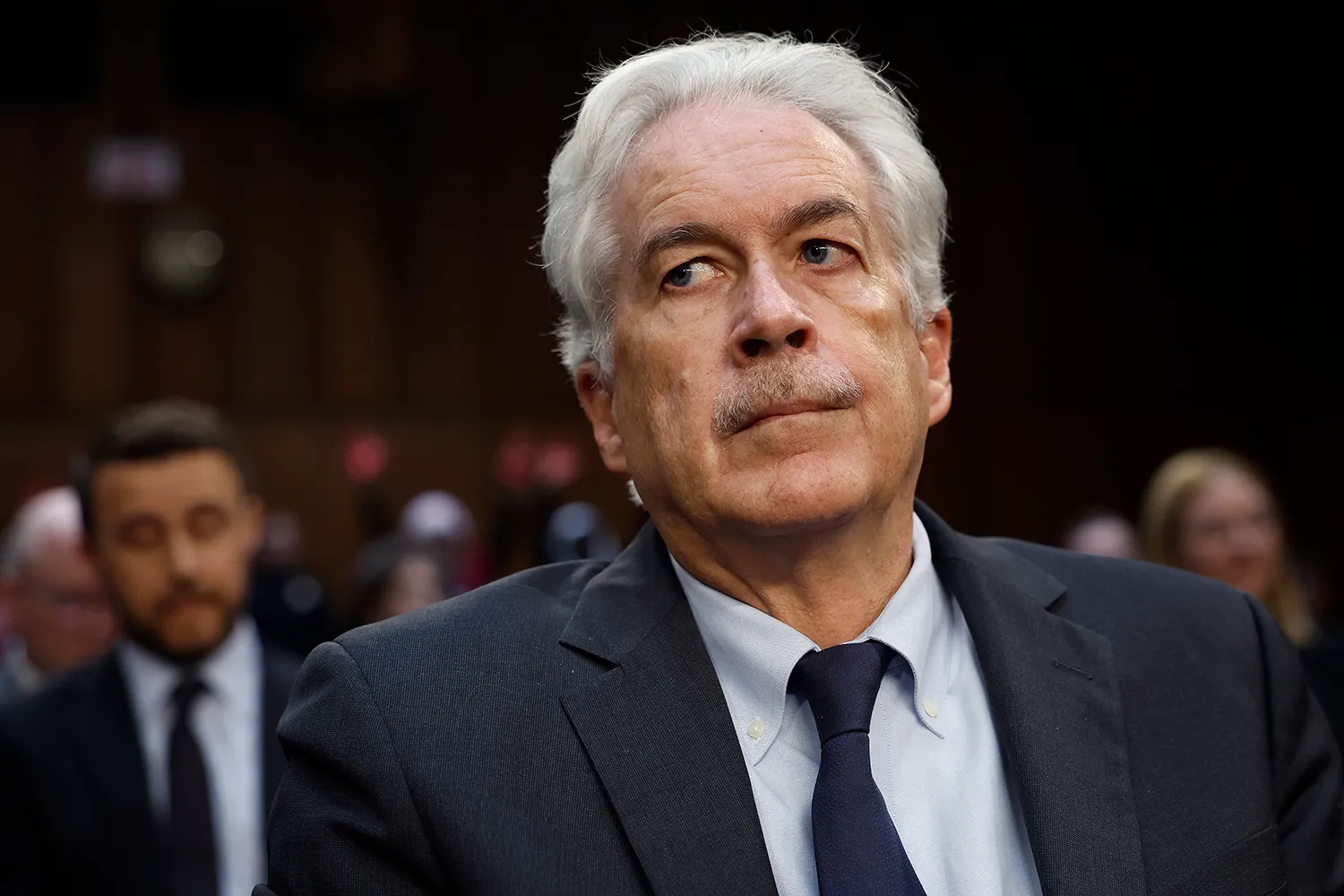 A man with white hair and a mustache wearing a suit looking serious with his eyes to the side.