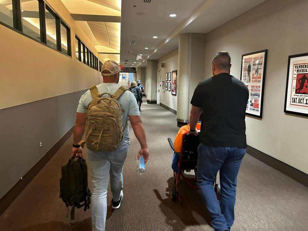 Two men walking down a hallway, one pushing a cart.