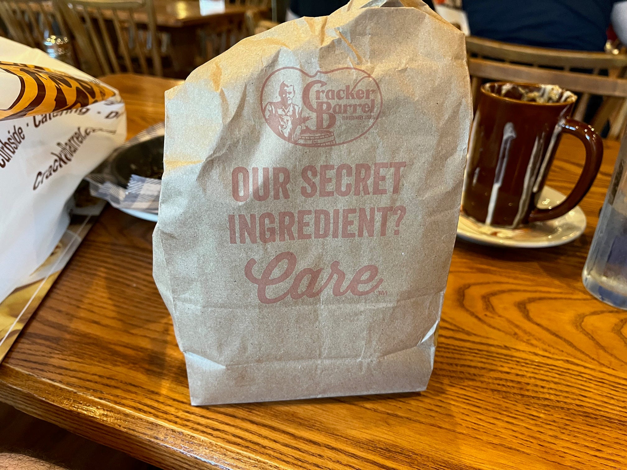 A brown paper bag that reads "Our Secret Ingredient? Care" sits on a table at a Cracker Barrel restaurant