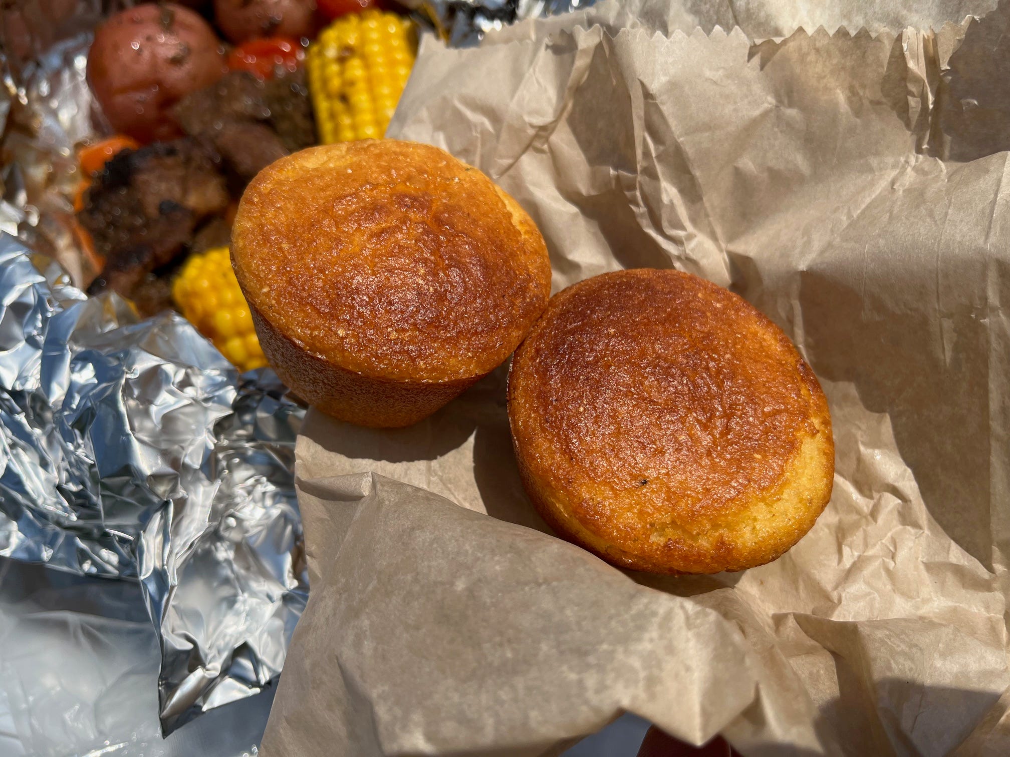 Two muffin-shaped pieces of cornbread sit on a paper bag with a beef campfire meal sits in the background.