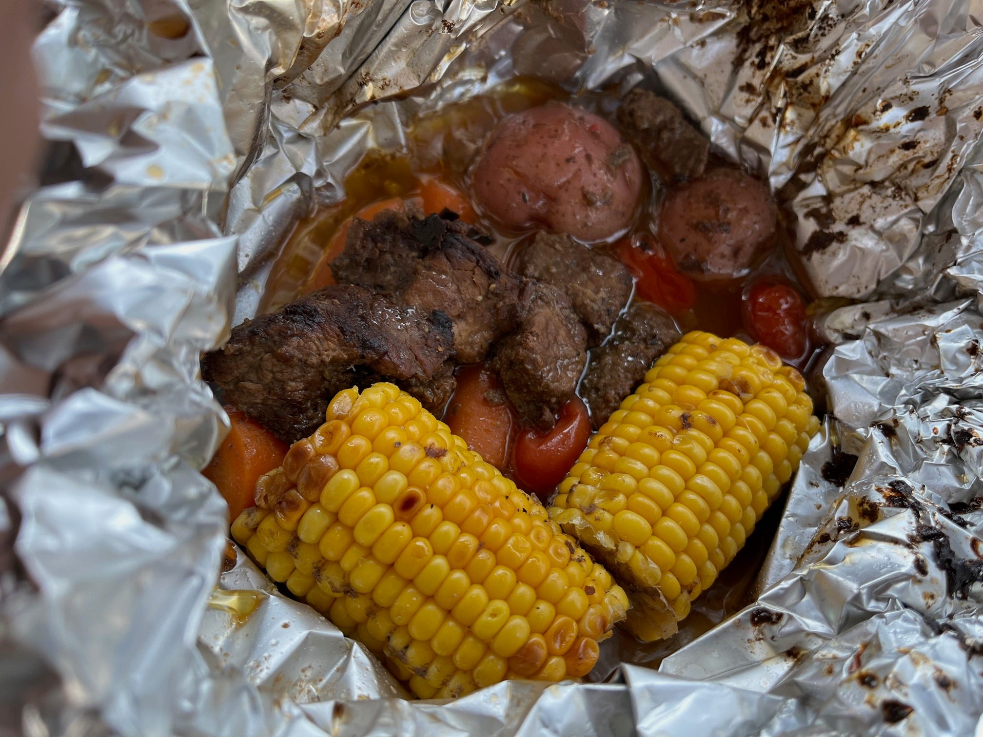A Campfire beef meal with potatoes, tomatoes, and two corn cobs wrapped in foil.