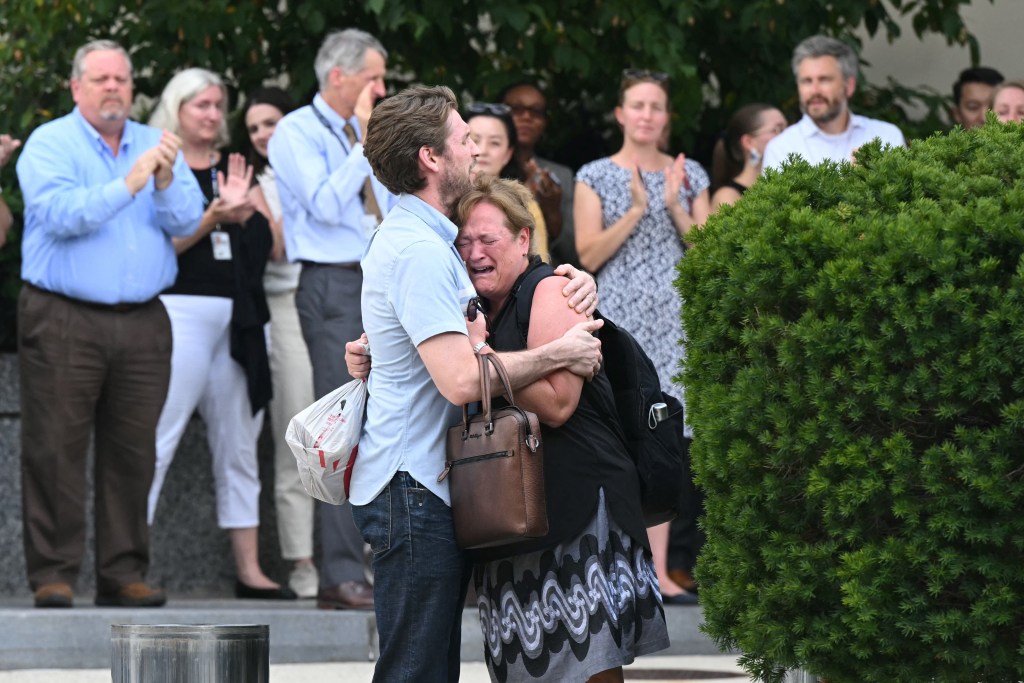 A tearful woman is comforted by a man as colleagues applaud nearby.