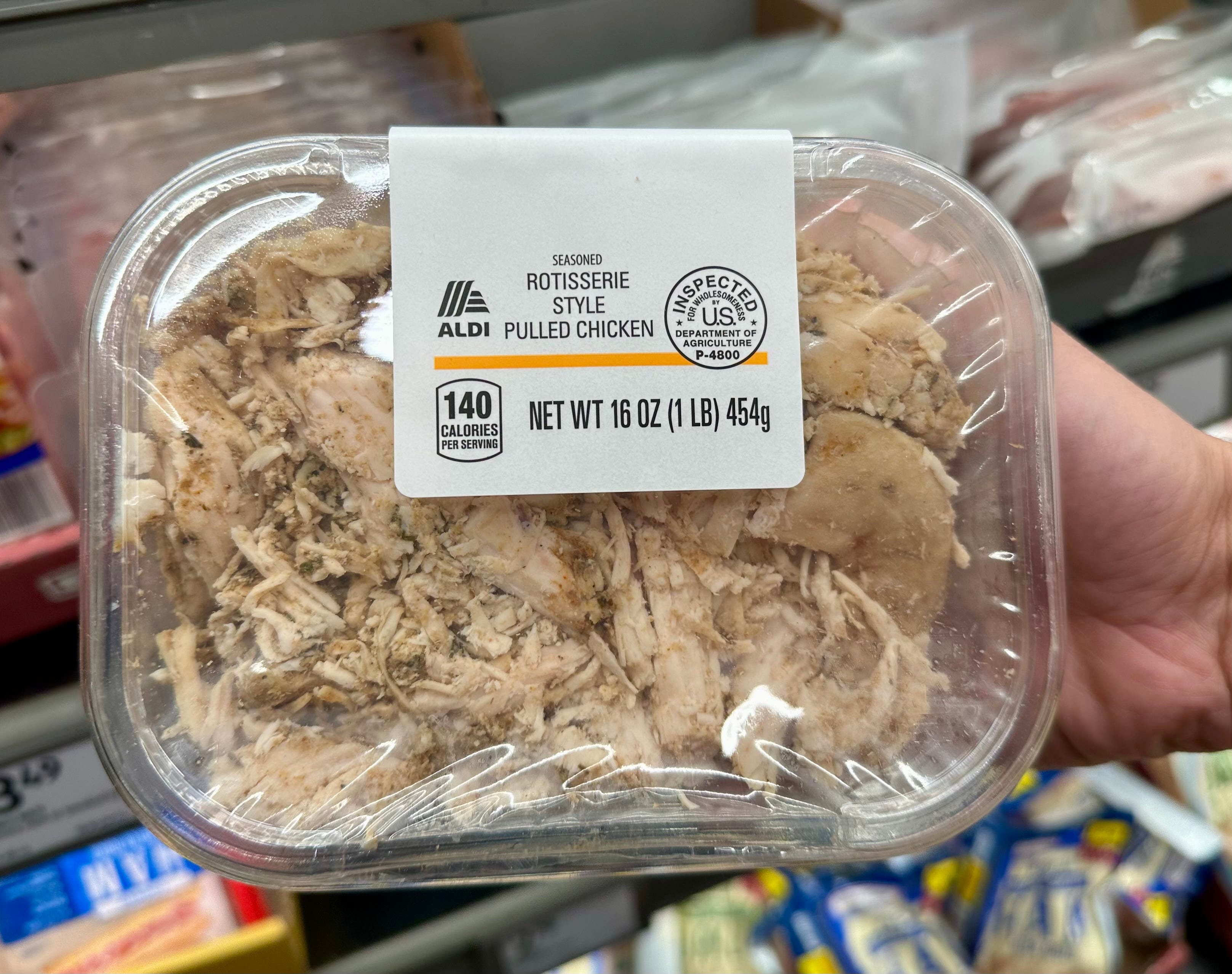 A hand holding a container of Aldi seasoned rotisserie-style pulled chicken.
