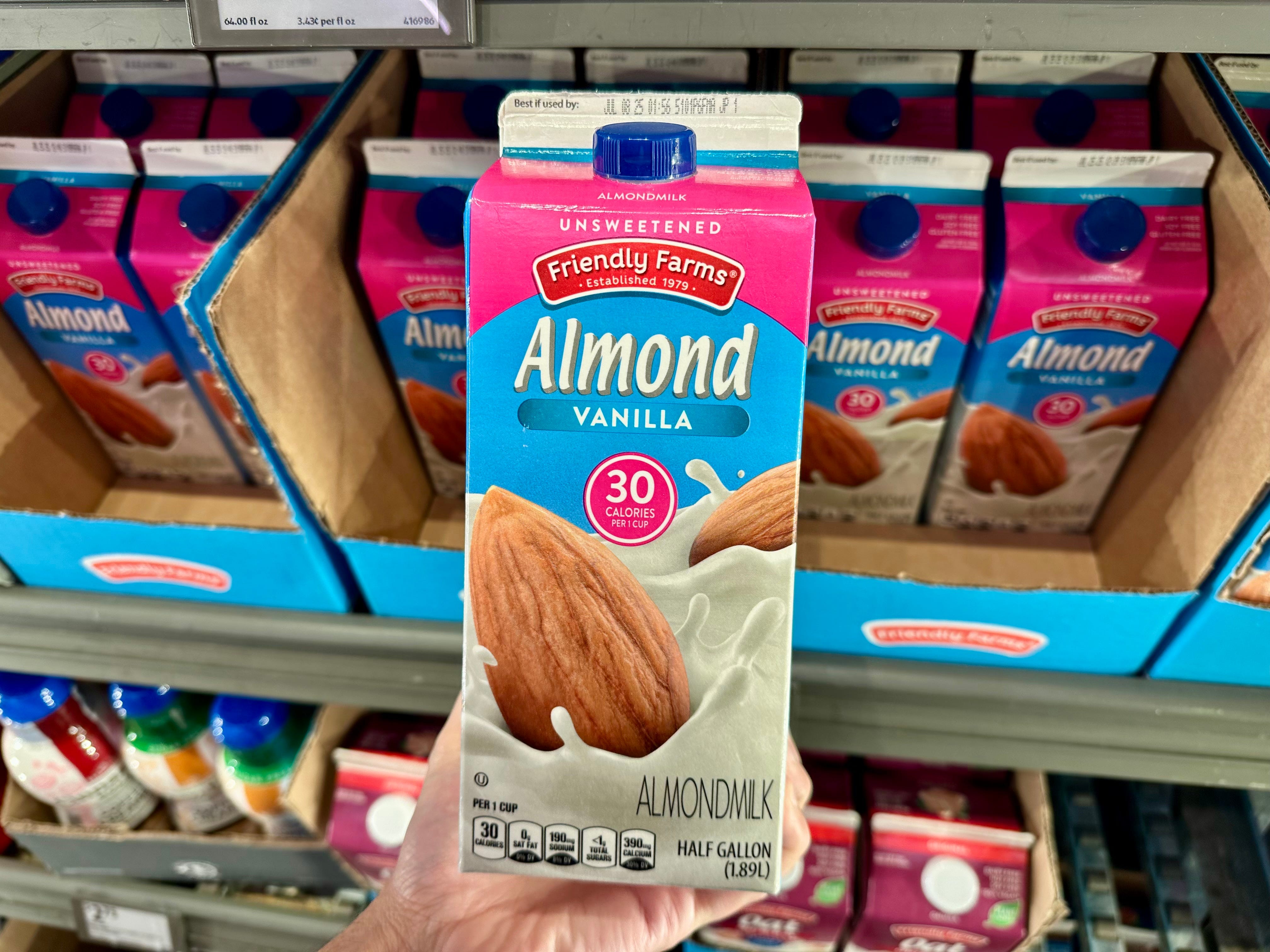 A hand holding a half-gallon carton of Friendly Farms unsweetened vanilla almond milk.
